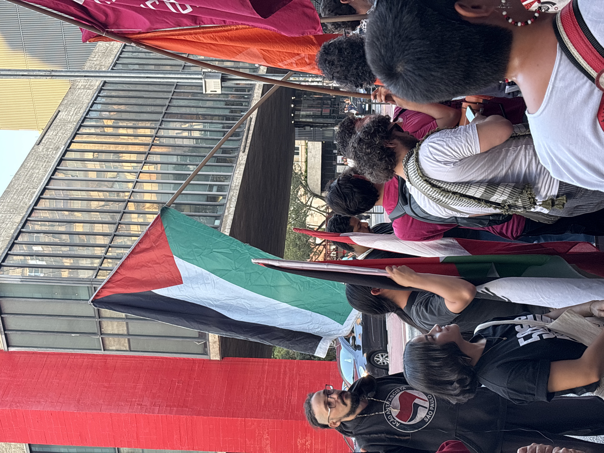 A medium, eye-level, outdoor shot depicts a street-level demonstration, likely in São Paulo, Brazil, as indicated by the Portuguese words "Ação Antifascista São Paulo" printed on a black hoodie worn by a man in the foreground.
The crowd, which is of mixed ethnicity, carries a Palestinian flag prominently in the frame. Red and burgundy banners are visible on the upper left side. The facade of a modern building is visible on the background. The windows are tinted with a metallic-reflective surface.
The man in the foreground, wearing a black hoodie, has dark hair with some graying in the sides and a dark beard. He also wears glasses and a beaded necklace. The woman next to him wears a black T-shirt with white lettering.
The demonstration seems to be focused and organized, with the participants holding flags and banners. The scene takes place during the day, with sunlight visible, reflecting on the glass of the building.