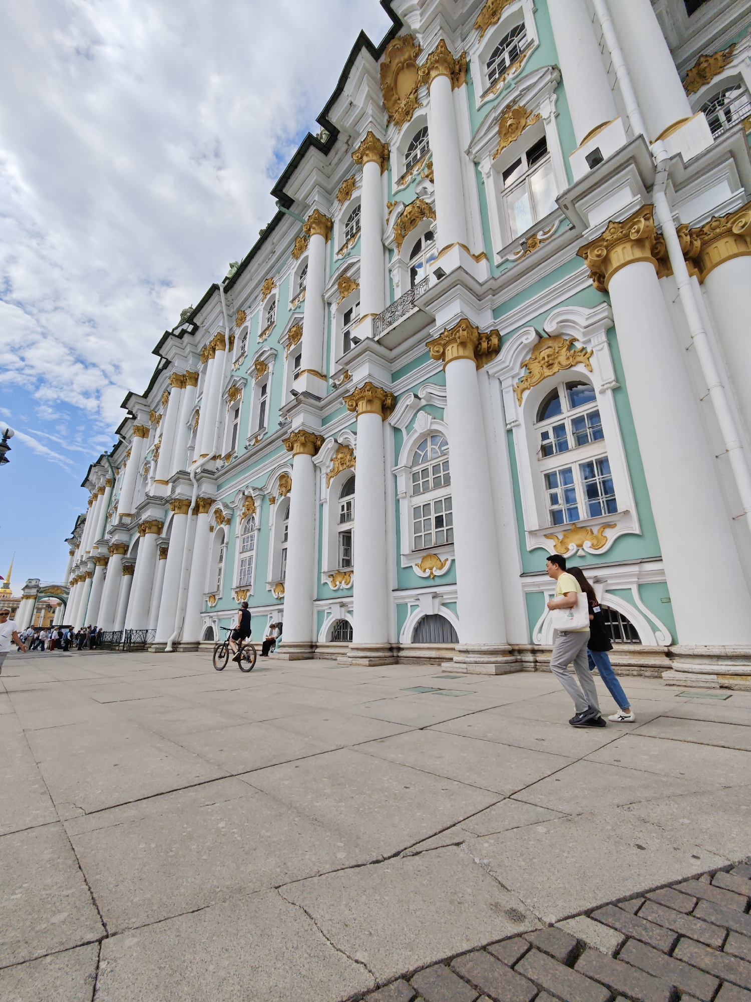 The image depicts a wide-angle, low-angle perspective of the exterior of the Winter Palace, a prominent landmark in Saint Petersburg, Russia. The palace facade is a striking light green with white and gold ornate detailing, featuring numerous tall, fluted columns topped with golden capitals. Multiple windows, some with arched tops, punctuate the facade.
In the foreground, a paved plaza extends towards the palace. On the plaza, a man is riding a bicycle from left to right across the frame. To the right of the cyclist, two people are walking from right to left. The man in front is wearing a yellow t-shirt and grey trousers, carrying a white tote bag. The person behind him wears a dark top and blue jeans. Further to the left, a crowd of people can be seen gathered, some appearing to be in a queue or observing the palace. In the far left background, a tall spire of another building is visible against a cloudy sky. The pavement is made of large, square concrete slabs, with some cracks visible. The sky is predominantly blue with scattered white clouds.