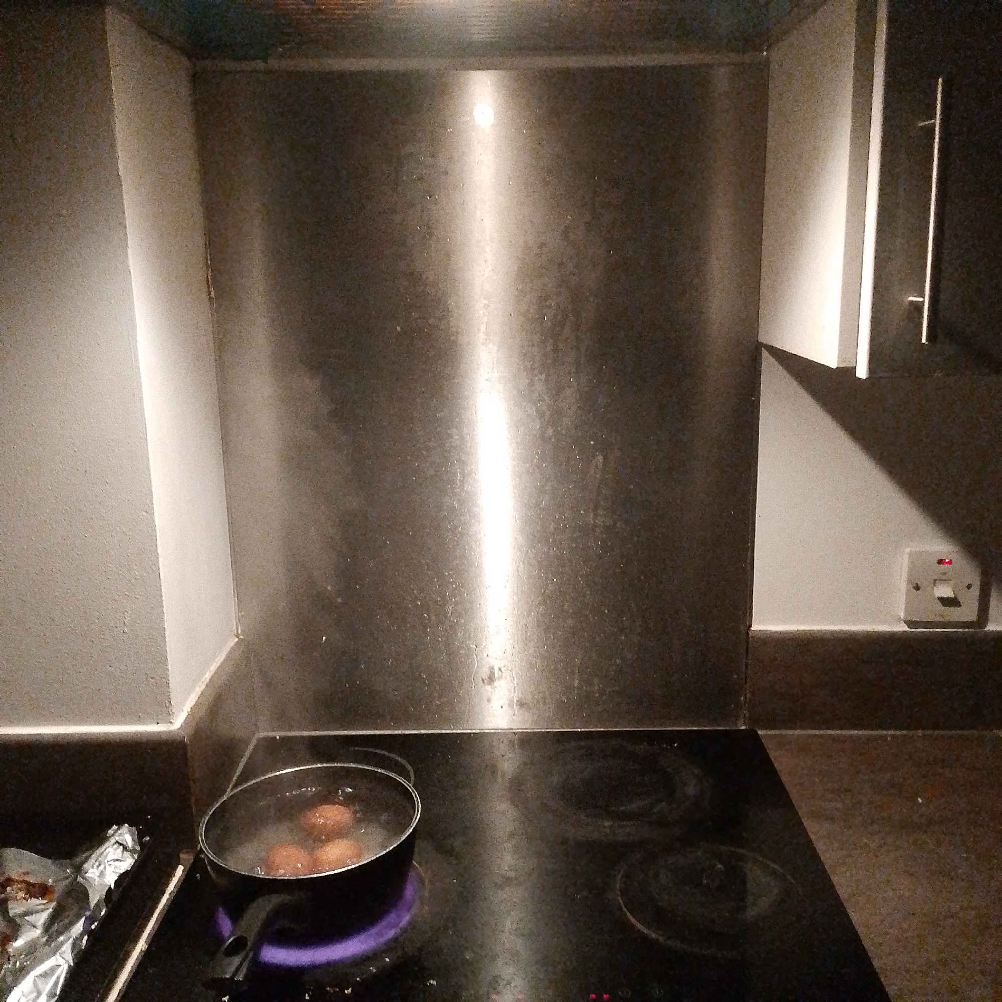 A dimly lit kitchen scene, likely in a residential dwelling, showing a black glass top electric stove with a pan of water and eggs boiling on the front left burner which is glowing purple. The stove is built into a dark countertop. To the left of the stove is a piece of aluminum foil with what appears to be cooked food remnants on it. Above the stove is a stainless-steel backsplash that reflects light. The walls on either side of the backsplash are painted a matte grey. To the right is a kitchen cabinet, dark in color with a silver handle, and a white electrical outlet is present on the wall next to the cabinet. Given the electrical outlet configuration, the location is most likely in the United Kingdom, possibly in Coventry, as requested. There are no people visible. The primary action is the boiling of eggs in a pan.