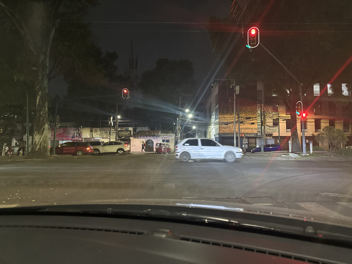 The image displays an urban street intersection at night in Mexico City, Mexico, viewed from inside a vehicle through its windshield, with the dashboard visible in the foreground. Three traffic signals are illuminated red: one overhead, one on the right, and another further down the street on the left. A white two-door hatchback, exhibiting motion blur on its wheels, is centrally positioned in the intersection, appearing to be in motion. To the left, a red sedan is stopped or parked behind a white SUV or crossover. On the far right, a red three-wheeled vehicle is parked adjacent to a single-story building with a distinctive arched entrance. A solitary individual stands near this building. Further down the street on the right, a dark blue or purple sedan is parked. Multiple multi-story buildings line the street, some with illuminated interiors. A large tree trunk is visible on the left side of the frame. Overhead utility wires, streetlights, and asphalt pavement are present, with the pavement appearing reflective under the ambient lighting conditions. A banner or sign above the parked red and white vehicles contains partially legible text including "BARRACO LA FINCA".