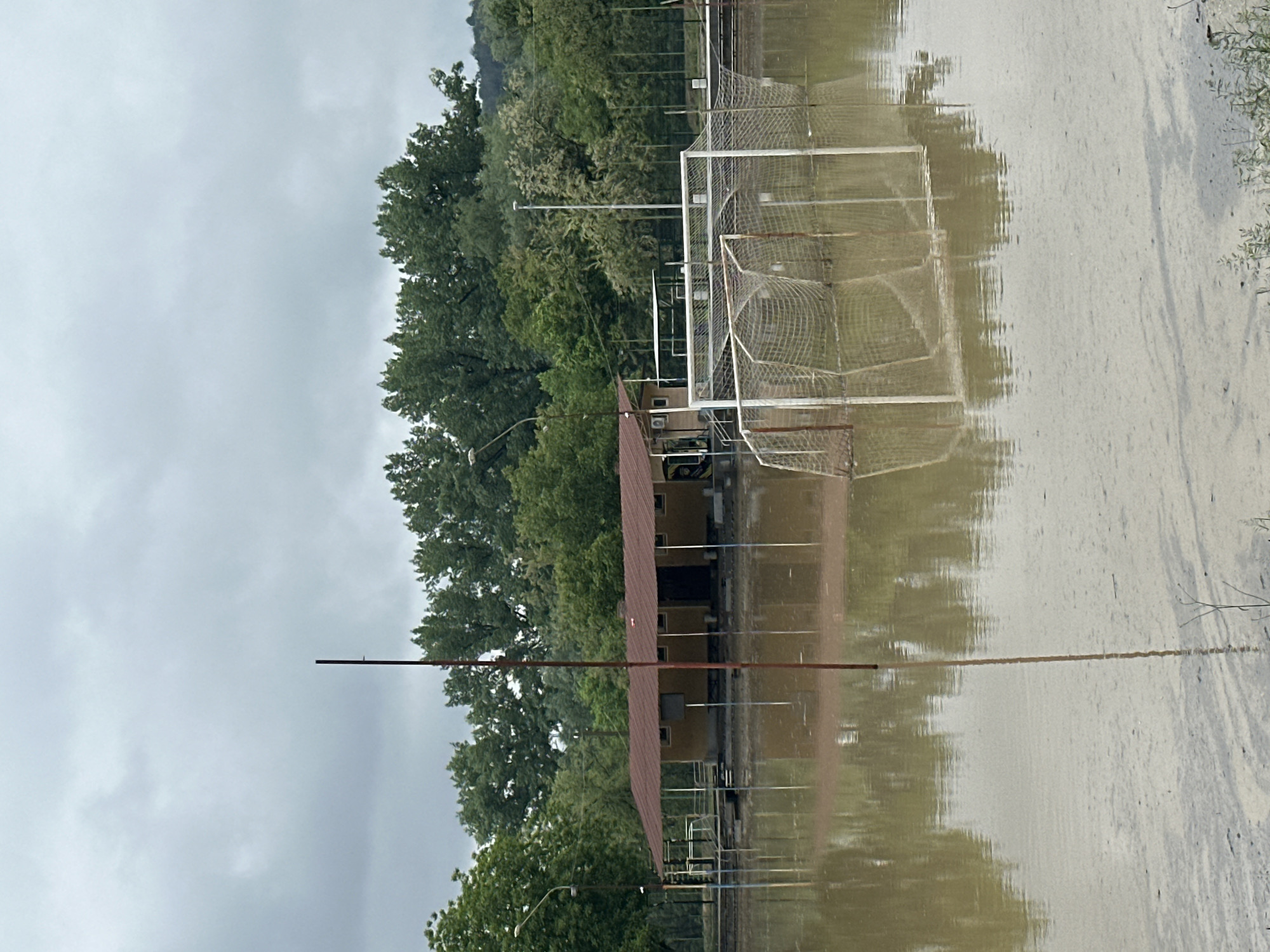 The image depicts a flooded soccer field, presumably in Debelets, Bulgaria, based on the description. Several soccer goals are visible, partially submerged in muddy water. A building with a red roof is situated next to the field, also appearing to be partially flooded. Trees are visible in the background, and the sky is overcast, suggesting recent rainfall. The water level is high enough to reflect the goals and building, indicating substantial flooding. No people are visible in the scene.