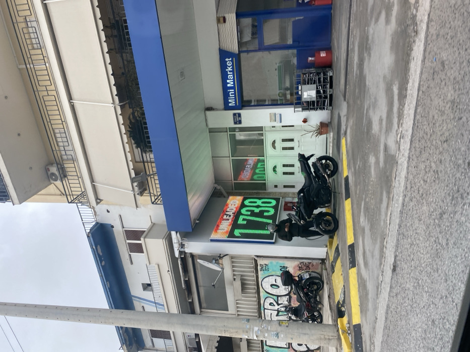 This image captures an urban street scene in Nikea, Greece, centered on a gas station with an attached mini-market on an overcast day. The gas station features a prominent blue canopy over the forecourt. A large digital LED sign, illuminated in bright green and red, displays "UNLEADED" and a price of "1.738," likely for fuel.
To the right of the price sign is the entrance to a "Mini Market," indicated by a blue sign above its glass storefront and a white, double-doored entrance. A red gas canister and a metal security cage are visible near the mini-market's entrance, along with a potted plant.
Two motorcycles are parked on the paved area in front of the gas station. A black motorcycle is positioned close to the price sign, with a person dressed in dark clothing standing beside it, possibly interacting with the vehicle or waiting. A second, smaller, dark-colored motorcycle is parked further down the sidewalk, nearer to a utility pole and a wall adorned with graffiti. The street curb alongside the gas station is painted with distinct yellow and black stripes.
In the background, behind the gas station, multi-story residential or commercial buildings are visible, featuring balconies and beige awnings. An air conditioning unit is mounted on one of these buildings, and a satellite dish is attached to the gas station building itself. A sturdy grey utility pole stands on the left side of the frame, extending towards an overcast sky.