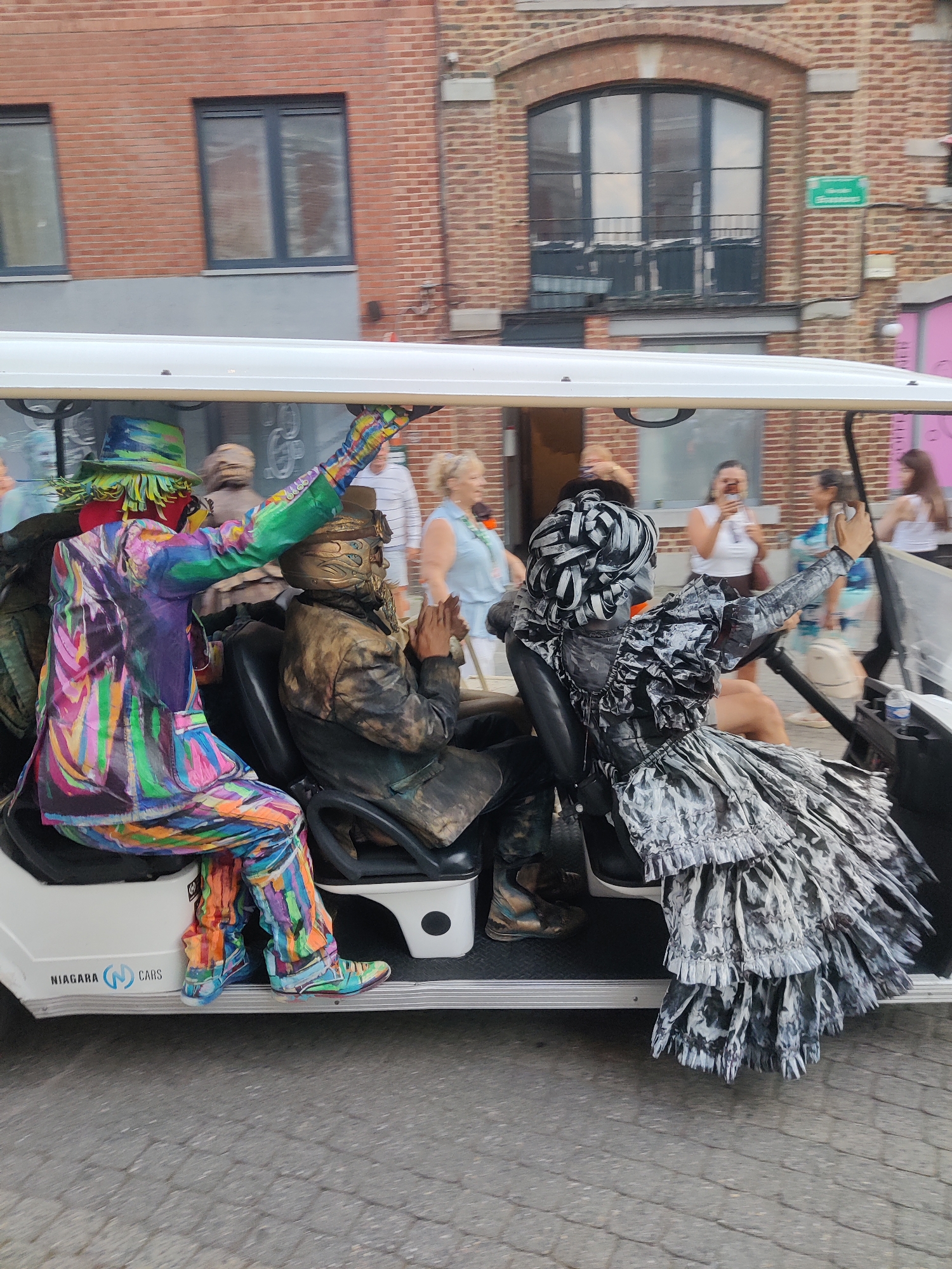 The image captures a street scene in Marche-en-Famenne, Belgium, with a white golf cart as the central object. Three individuals, seemingly dressed in elaborate costumes or body paint, are seated within the cart. The person in the front left is wearing a vibrant, multi-colored outfit with a matching hat and has their arm raised. To their right, another individual is adorned in a metallic, possibly bronze-colored costume that covers their head and body, with their hands clasped. The person in the back right is dressed in a black and white, ruffled costume resembling a voluminous dress and has their arm extended, holding what appears to be a cell phone. The cart has "NIAGARA CARS" branding on its side. In the background, a brick building with arched windows is visible, suggesting an urban or town setting. Other people are blurred in the background, suggesting movement and activity. The overall impression is of a public event or parade taking place on a paved street.