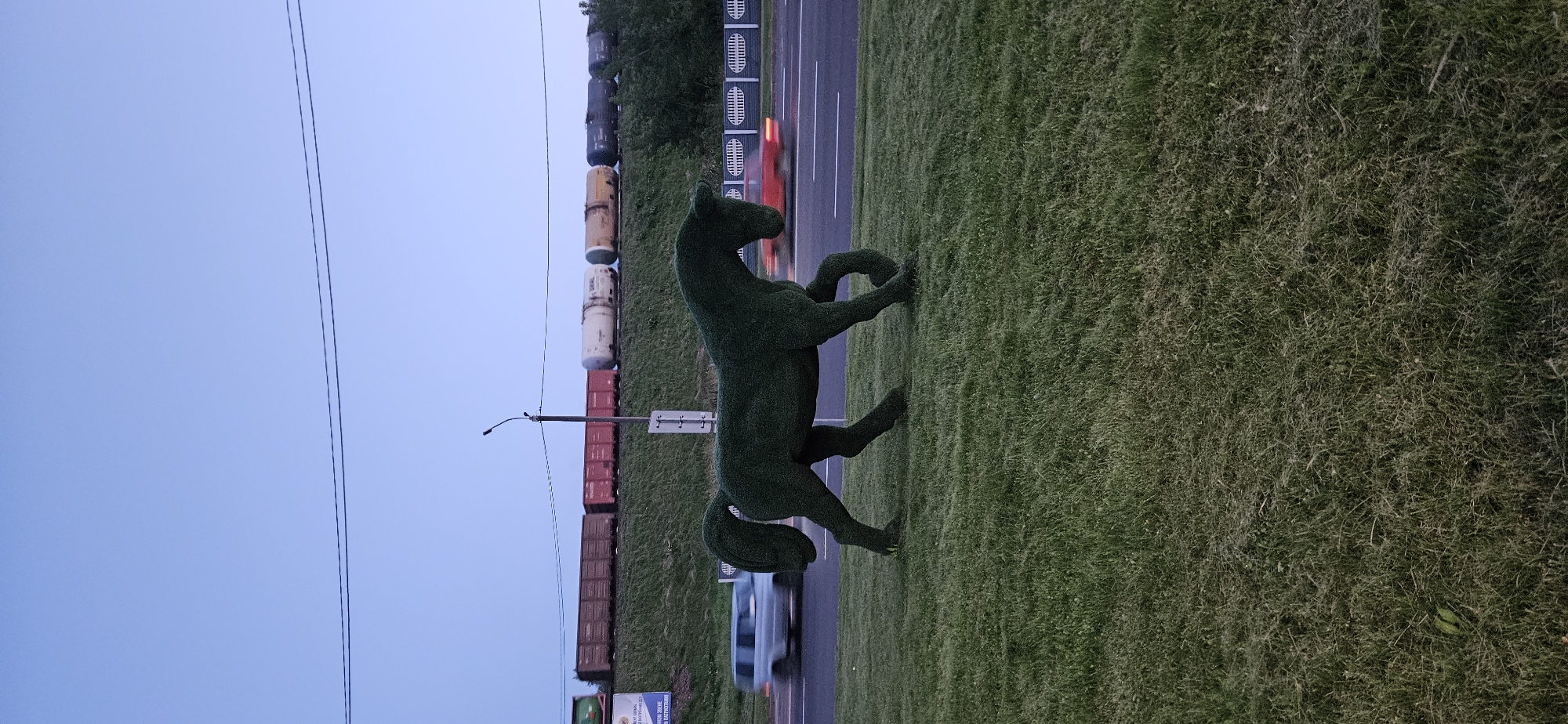 The image shows a topiary sculpture of a horse standing on a grassy area next to a road. In the background, a train with several carriages is visible on elevated tracks, and a billboard with Russian text is partially visible. Cars are seen driving on the road, with motion blur indicating their movement. The sky is a light blue, suggesting dusk or dawn. The scene is located in Vitsebsk, Belarus, on the side of a road near a railway line.