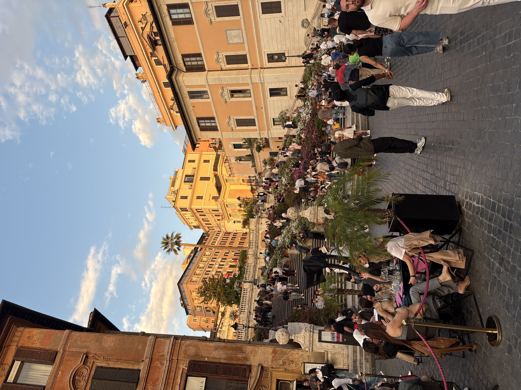 The image depicts the Spanish Steps in Rome, Italy. The scene is bustling with people sitting on the steps, socializing, and enjoying the surroundings. The steps are decorated with plants and flowers. Buildings line the sides of the steps, showcasing varied architectural styles and colors, ranging from warm oranges and yellows to muted browns. The sky is partially visible with scattered clouds. In the foreground, there's a roped-off area with tables and chairs, suggesting an outdoor cafe or dining area. People are seated at the tables, and there are elements like glassware and decorative plants. The cobblestone pavement adds to the urban environment.