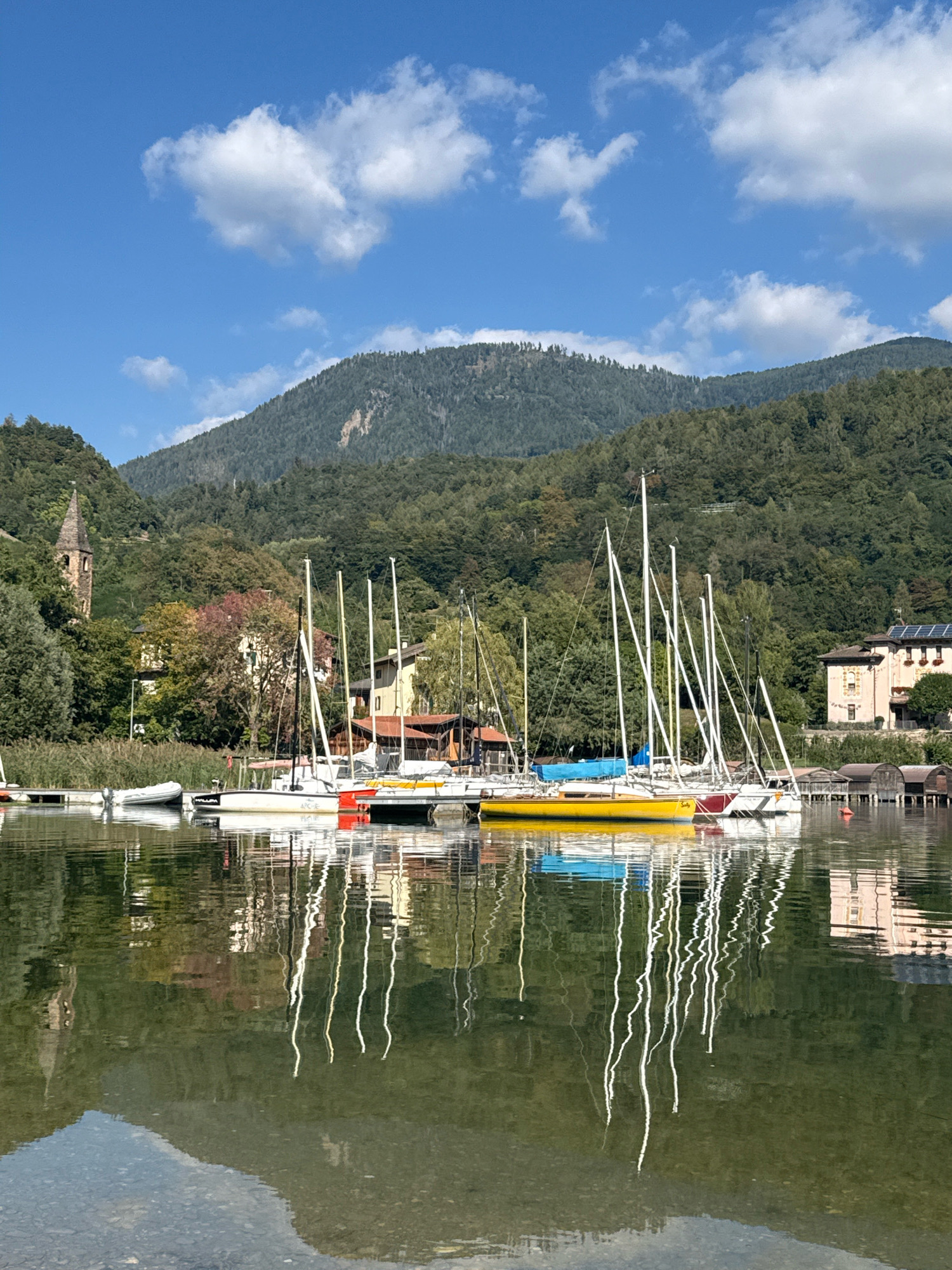 A sunny, bright day features a calm lake with several sailboats docked at a wooden pier. The boats, varying in size and color, have tall masts that extend into the clear blue sky. The lake's surface is a mirror, reflecting the boats, their masts, and the surrounding lush green forested mountains and buildings.
On the left side of the image, a tall, old church tower with a pointed spire is visible amongst trees. Further to the right, residential buildings with terracotta roofs and tan facades are nestled into the hillside. The water in the foreground is clear, revealing a rocky bottom. The image captures a peaceful, idyllic scene in Pergine Valsugana, Italy.