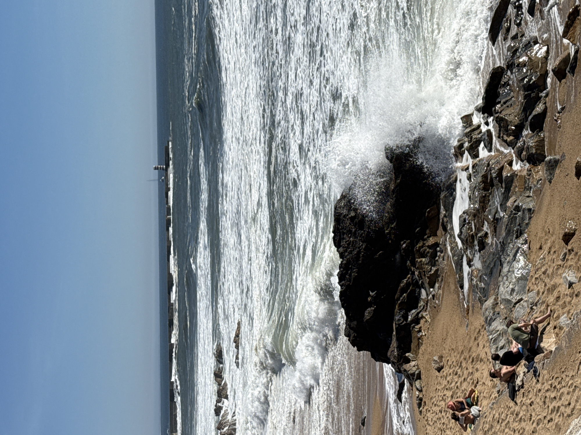 The image shows a beach scene in Porto, Portugal, featuring a large rock formation where waves are crashing. The sky is clear and blue. In the distance, a pier with a lighthouse can be seen. In the foreground, there are three people relaxing on the sand near the rocks. One person is lying on their back, another is lying on their stomach, and the third is partially obscured but appears to be in a similar relaxed pose. The beach appears to be sandy with darker rocks and stones scattered along the shoreline. The dominant action is the waves hitting the rocks, creating white foam and spray.