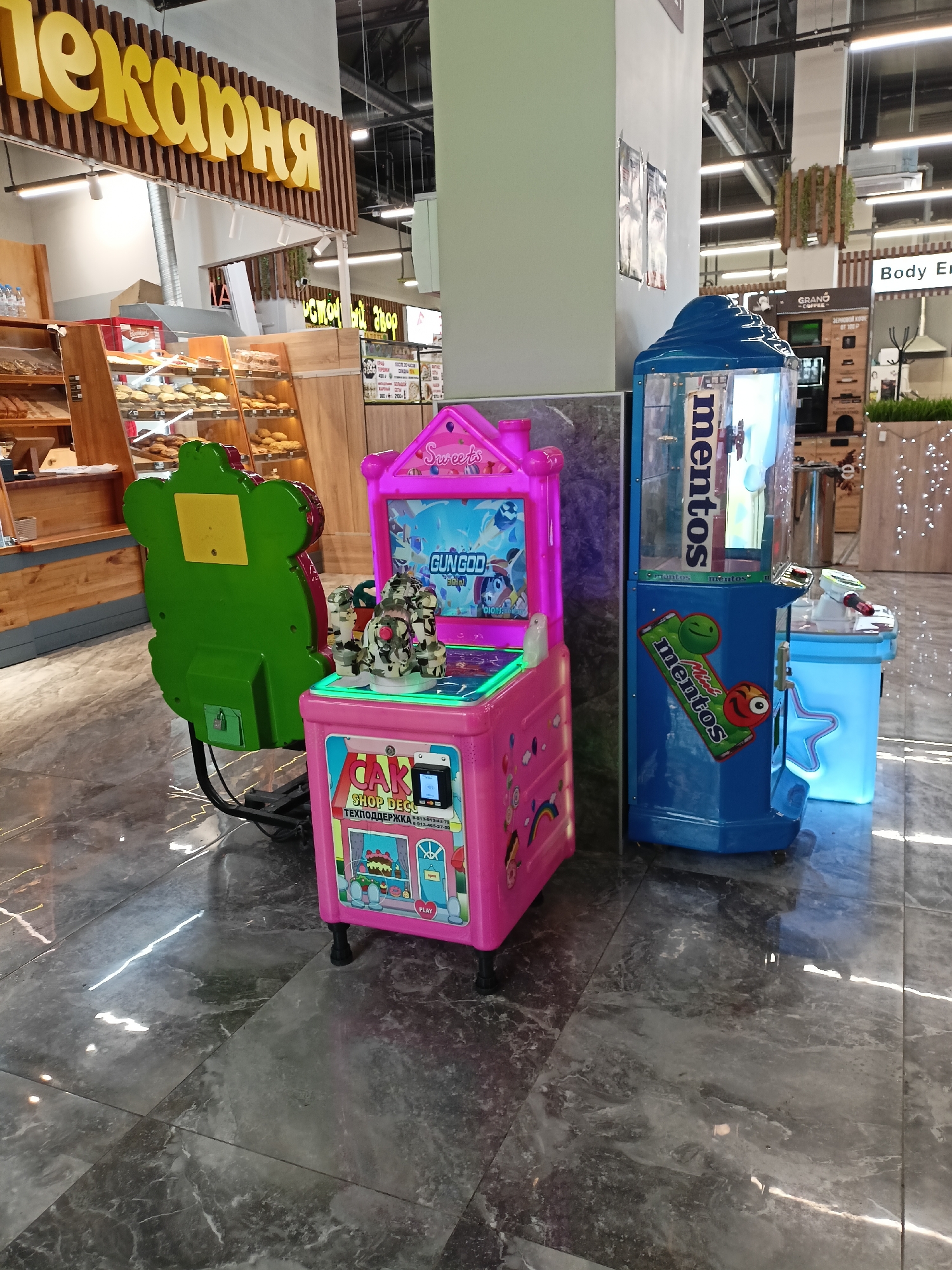The image depicts a brightly lit, modern shopping mall in Novosibirsk, Russia, featuring a mix of retail establishments. In the foreground are three colorful arcade-style game machines: one pink with a "GUNGOD" game screen, one shaped like a green bear, and one blue with "mentos" branding. The game machines are standing on a shiny gray-tiled floor. A bakery ("Пекарня" in Russian) is visible in the background, displaying a variety of baked goods. In the distance, there is a "Body E" brand advertisement and other retail store displays, suggesting a diverse range of shops within the mall. There are no people visible in the immediate frame. The scene is well-lit and orderly, indicating a clean and well-maintained shopping environment.