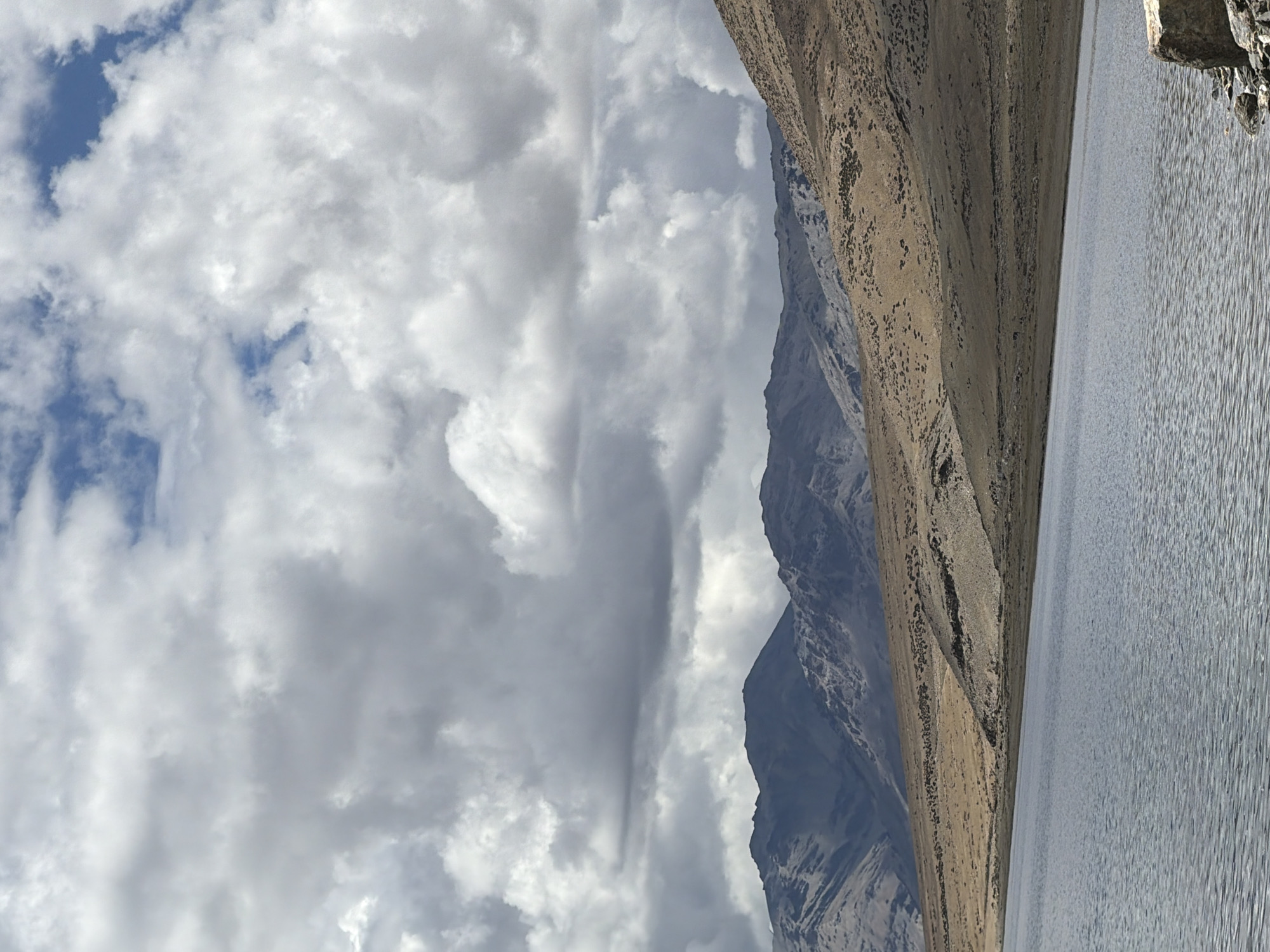 This is a vertical, eye-level shot of a vast, serene lake bordered by barren, rugged hills under a dramatic, cloudy sky. The lake's surface is a muted blue-grey with subtle ripples, reflecting the overcast heavens. The shoreline transitions from a dark, rocky band to a tan, dusty slope dotted with sparse vegetation, which in turn gives way to towering, rocky mountains in the distance. These mountains are layered with shadows and light, revealing their textured, uneven surfaces. The sky is filled with a dense accumulation of grey and white clouds, with patches of bright blue peeking through, suggesting an intermittent sun. The overall environment is wild and uninhabited. The bottom right corner shows a few rocks along the lake's edge.