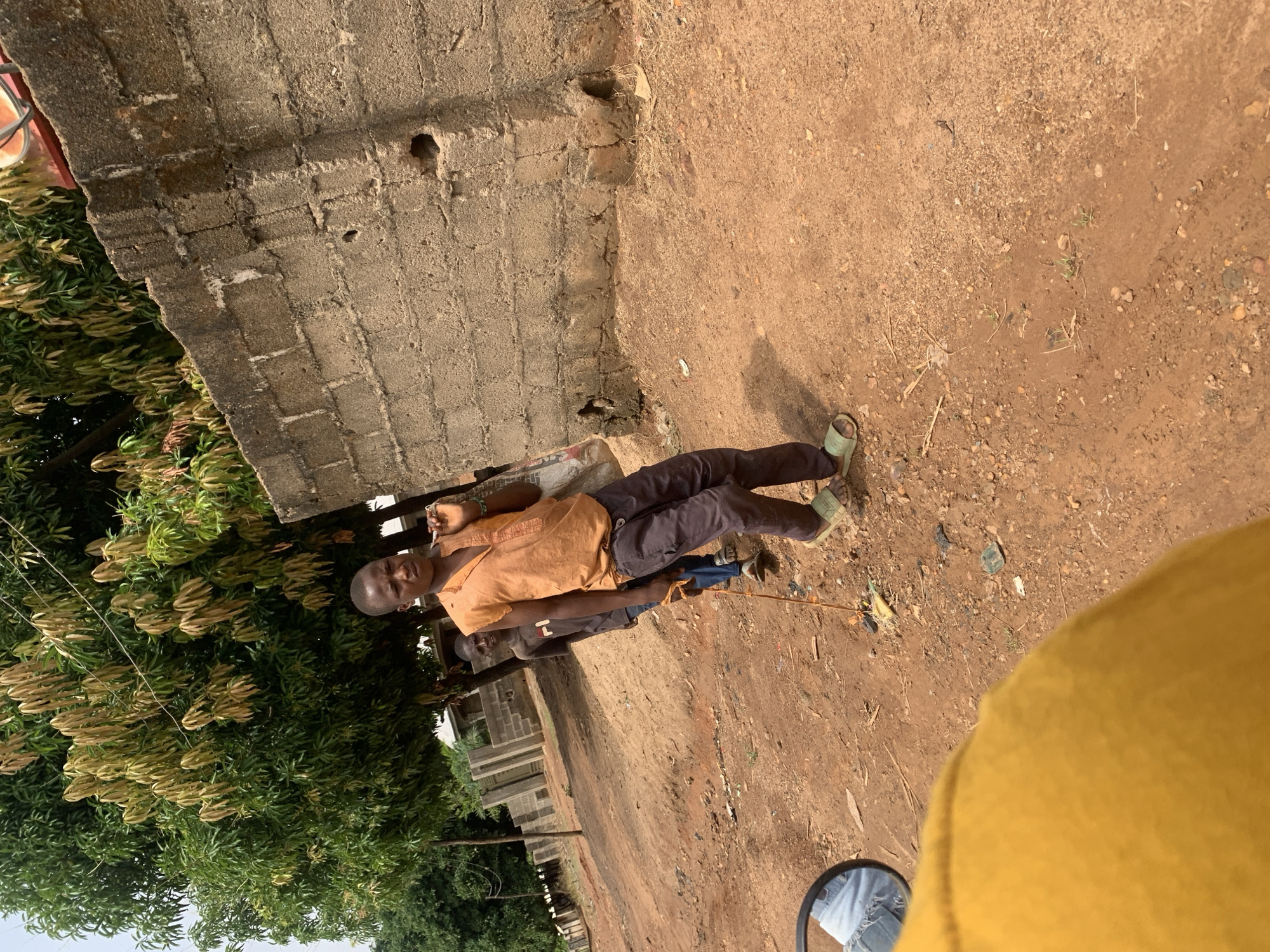 The image depicts two individuals outdoors in what appears to be a street scene in Akwanga, Nigeria. A young man, dressed in an orange shirt and dark pants, is reclining on a padded surface on the ground, holding a slender stick-like object. Another person is lying on the ground slightly behind him in the same direction and is wearing dark clothing. There are trees and vegetation along the left edge of the frame, and a concrete or brick wall is visible along the top left. The ground is dirt or reddish-brown soil with scattered debris. A part of a yellow object and a rearview mirror are visible in the foreground, likely part of the vehicle from which the picture was taken. The buildings in the background appear to be under construction.