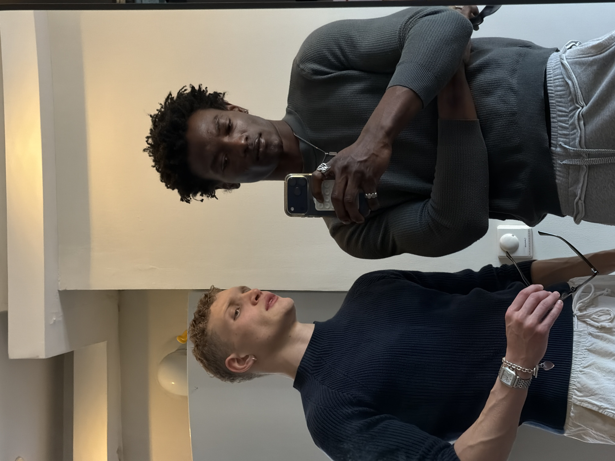 Two men are taking a mirror selfie. One man has dark skin, dark curly hair, and is wearing a gray sweater and gray sweatpants. He is holding a phone in his hand, and there is a silver necklace around his neck. The other man has light skin, short blond hair, and is wearing a dark blue sweater and white pants. He is holding a pair of glasses in his hand, and there is a silver watch and bracelet on his wrist. The location appears to be an indoor setting with white walls. There is a light fixture visible. It is not possible to determine whether this is in London, UK, from the information present in the image.