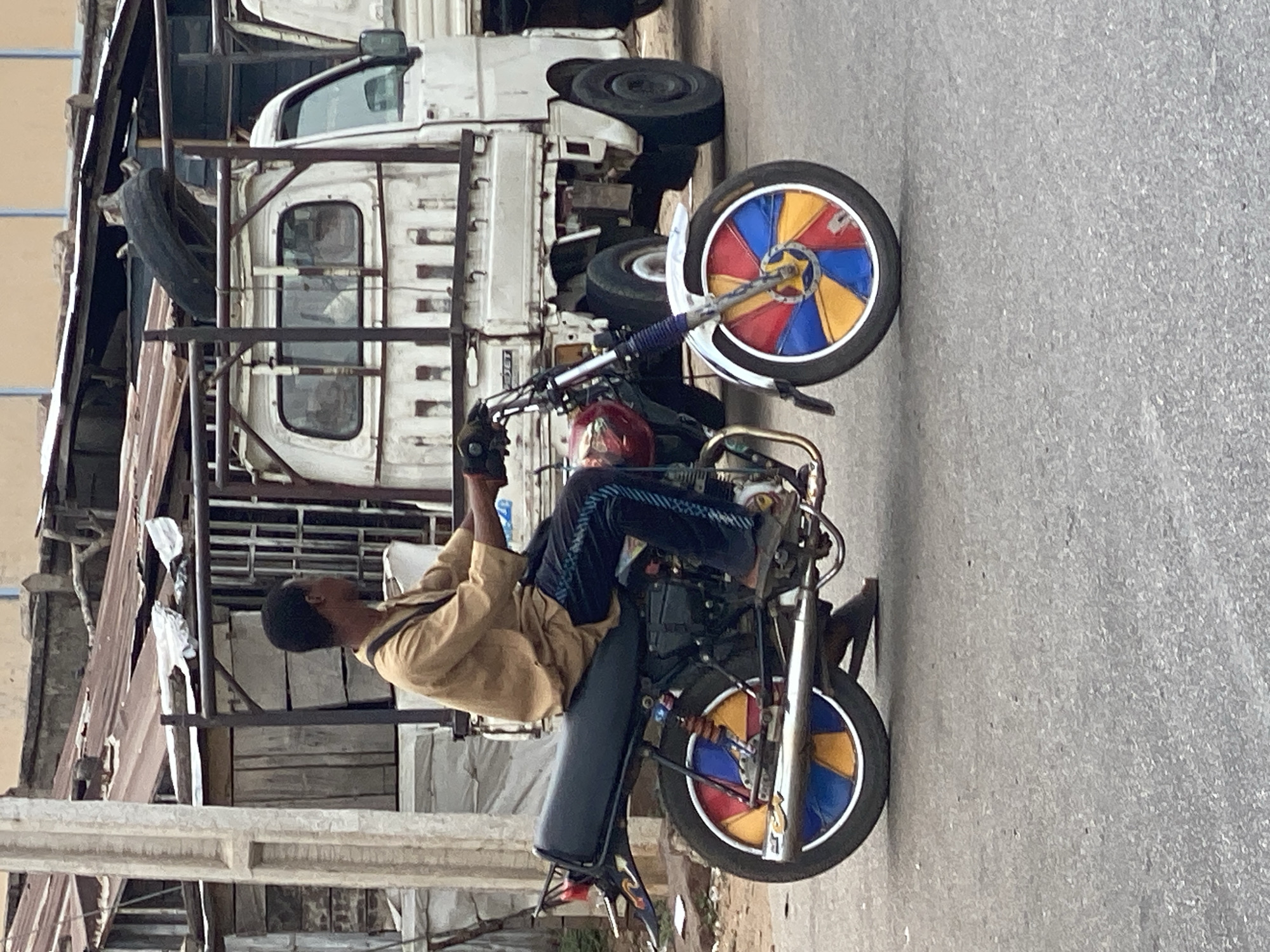 The image depicts an outdoor urban or semi-urban scene, likely in Lokoja, Nigeria, during daylight. The central subject is a young Black man riding a black motorcycle, performing a wheelie with the front wheel elevated high off the ground. He wears a light brown or tan long-sleeved shirt and dark blue pants with light-colored stripes. His posture suggests concentration and balance as he leans back, controlling the motorcycle.
The motorcycle is highly customized, notably by its wheels. Both the front and rear wheels feature a distinctive, brightly colored pinwheel design, divided into segments of red, yellow, and blue. The background is dominated by several white pickup trucks or small utility vehicles, some appearing worn and stacked. Metal scaffolding or framework, along with parts of industrial or dilapidated structures, further characterize the setting. The ground is paved, likely asphalt or concrete. The scene conveys a sense of everyday life, possibly a street or an area near workshops, with the motorcycle rider adding a dynamic element. No readable text is visible.