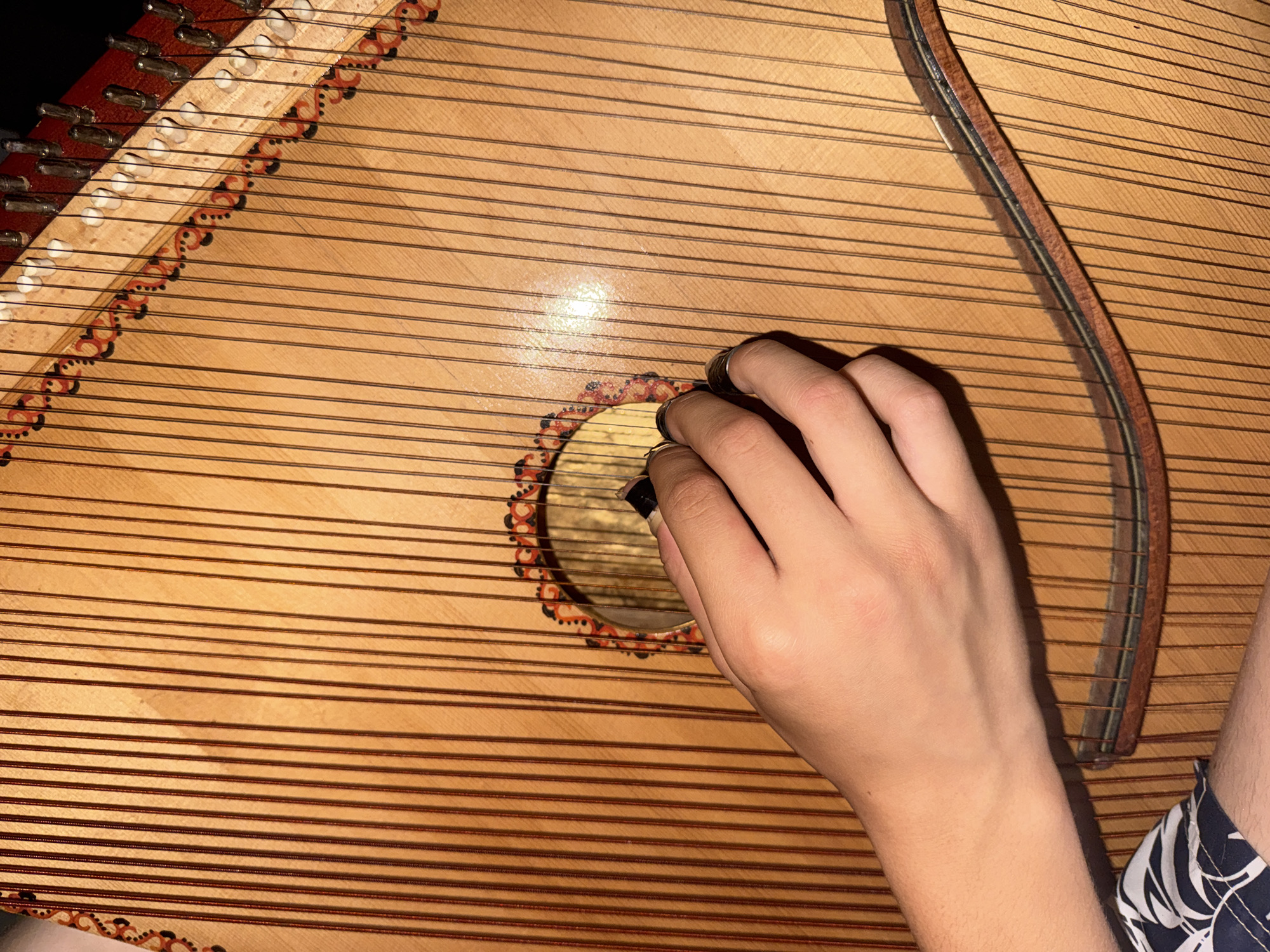 A close-up, eye-level shot shows a person's hands interacting with a bandura, a Ukrainian plucked string instrument. The instrument's light-colored wooden body is covered in numerous strings. A hand with dark-painted fingernails is visible on the right, with fingers positioned over the strings near the soundhole. The left arm and hand are also partially visible in the lower left of the frame, resting on the instrument. The environment is dark, with light reflecting off the instrument's surface.