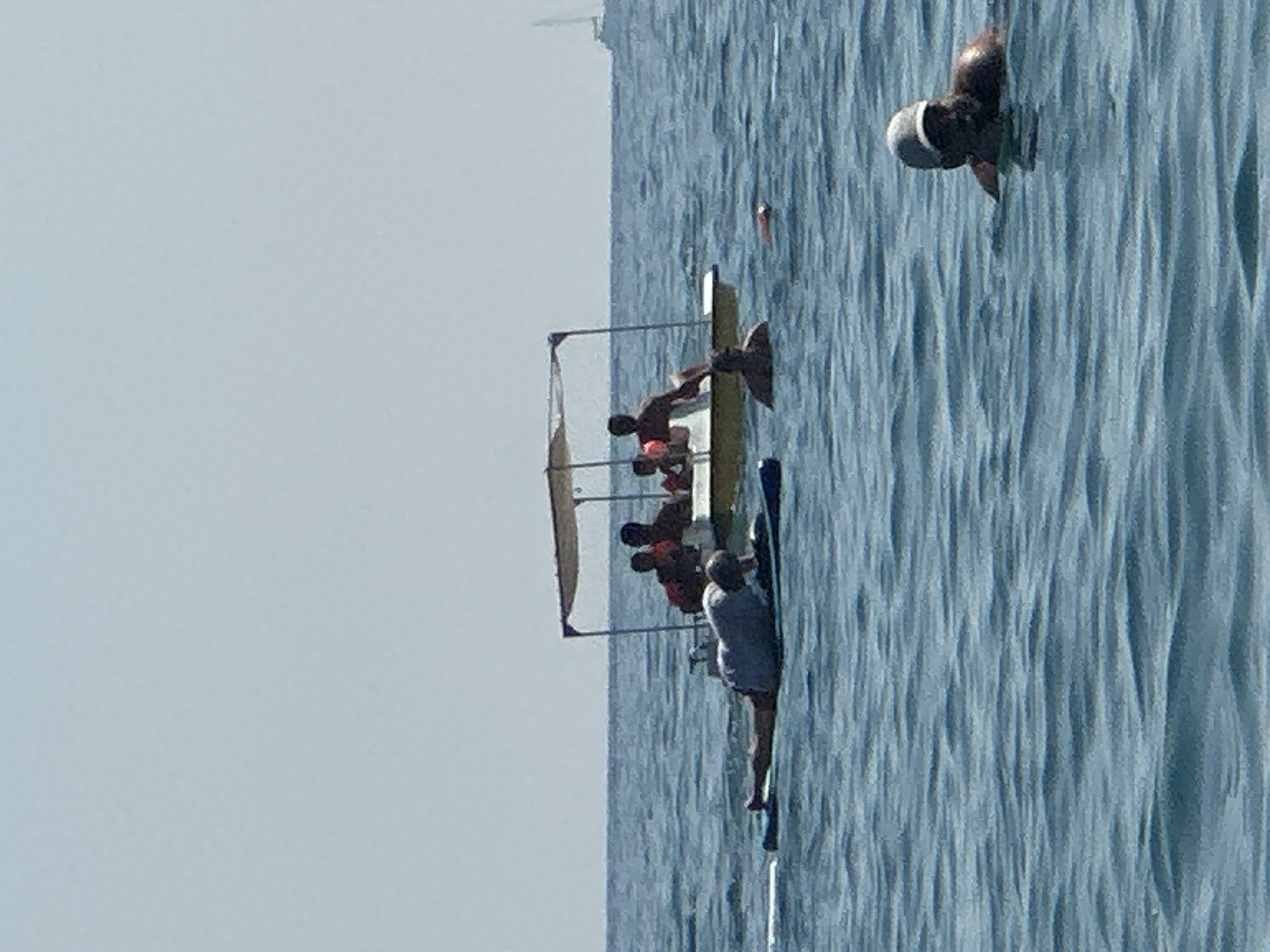 In the coastal waters of Boreti, Montenegro, a group of individuals are engaged in various aquatic activities. In the foreground, a person wearing a cap is submerged in the water, with only their head and shoulders visible. To the left, another individual is lying prone on a paddleboard, seemingly resting or observing the surroundings. Further out in the water, a small boat with a canopy is carrying several people. Some are seated, while others appear to be standing or moving within the vessel. The water is a tranquil blue, with gentle ripples suggesting a calm sea. In the distance, a sailboat can be spotted on the horizon under a clear, pale sky. The scene captures a leisurely day at the beach with recreational water sports and boating.
