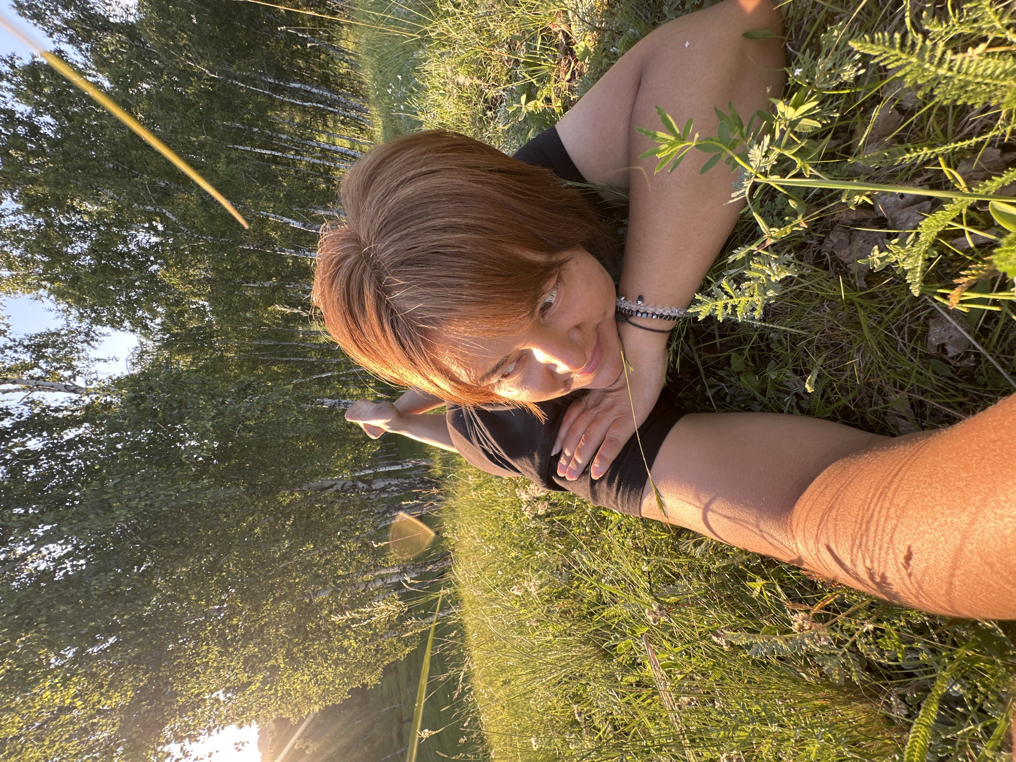 A woman with auburn hair lies in the grass, her face tilted towards the camera, a gentle smile on her lips. She is wearing a black top and black shorts. Her left arm is bent, with her hand resting near her face, while her right arm is extended behind her head. Her legs are bent at the knees and spread apart, suggesting a relaxed or acrobatic pose.
The setting is a grassy field with a line of trees in the background. The trees appear to be birch trees, with their distinctive white bark. The sun is low in the sky, casting a warm, golden light on the scene and creating long shadows. This suggests that it is either early morning or late afternoon. The weather appears to be clear and sunny. There are some small white flowers scattered in the grass. The overall mood is peaceful and serene. There is no visible text in the image.