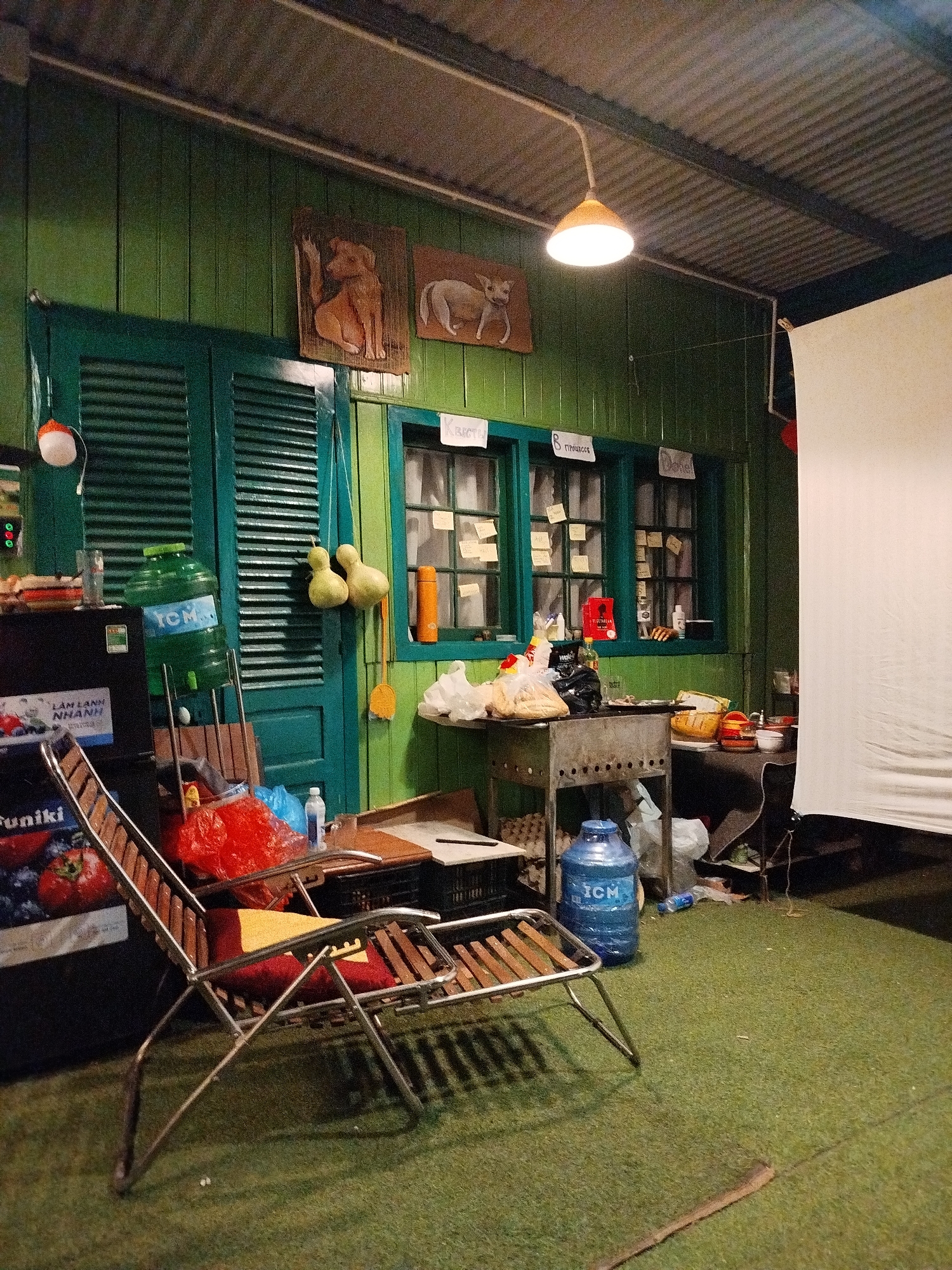 The scene appears to be inside a rustic building in Cam Ly - Da Lat, Vietnam. The walls are green painted wood paneling. A double door and several windows are also painted green and adorned with handwritten notes. Two pieces of art depicting animals hang above the door. A hanging light illuminates the center of the room. Various items are scattered throughout, including bottles, bags of food, and a water cooler. A metal grill sits near a table laden with dishes. A folding lounge chair with a red and yellow cushion is placed in the foreground. A white backdrop covers the right portion of the scene. The floor is covered in what appears to be artificial grass.