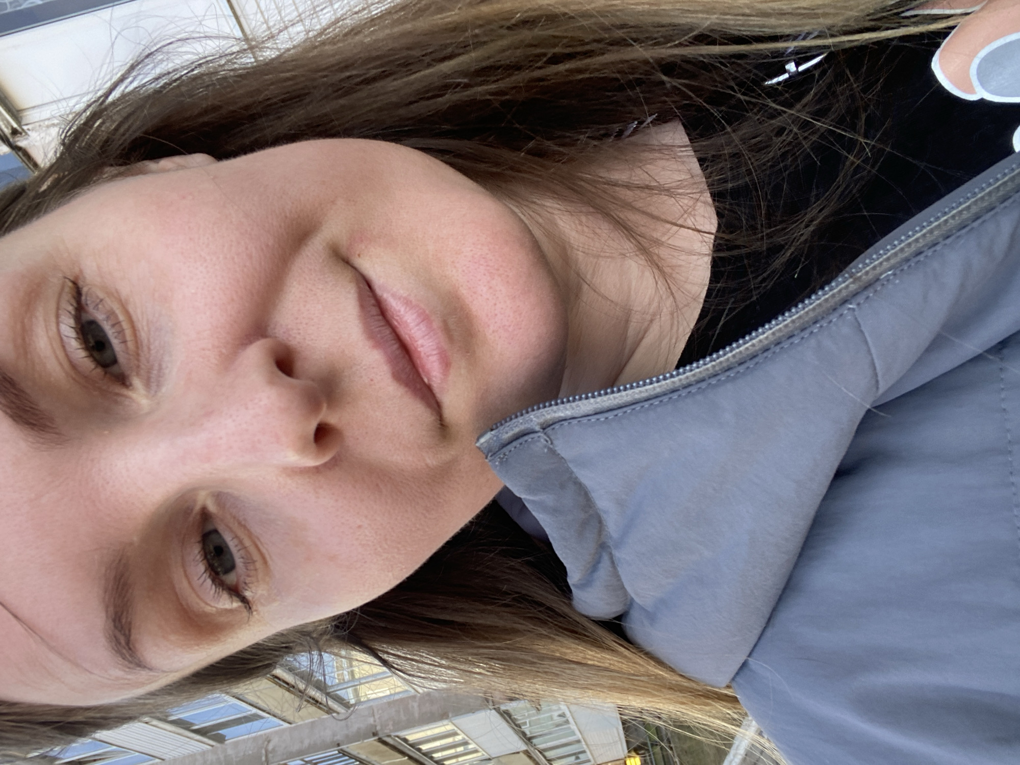 A young, fair-skinned woman with light brown hair is shown in a close-up selfie-style image. She has a slight smile and is wearing a black t-shirt with a white graphic design, partly visible, and a light blue jacket. The background appears to be a blurred urban setting with architectural elements resembling a glass-roofed walkway or building, possibly a transit station. The location is Noviy, Russia.