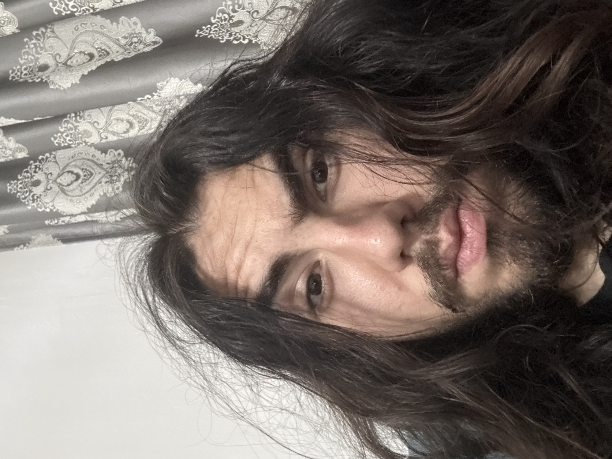 An extreme close-up selfie shows the face of a person with long, dark, unkempt hair and a goatee. The person's eyes are open and looking at the camera. The background shows a textured grey curtain with a damask pattern and a plain white wall.