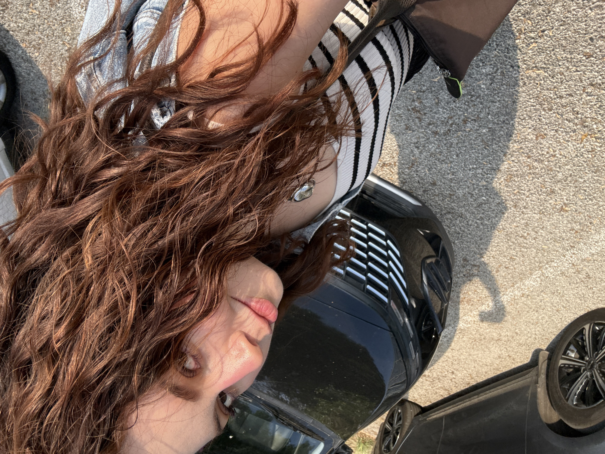 The image displays a young woman posing outdoors, seen from a slightly high and tilted angle. Her long, wavy brown hair, cascading over her shoulder and chest, is a prominent feature. She is wearing a light-colored top with black horizontal stripes on the upper arm/shoulder area, layered over what appears to be a white tank top, and light-wash denim is visible on her lower body. A silver-colored pendant hangs around her neck. Her face, seen in profile looking towards the camera, shows light makeup, distinct eyelashes, and pink-tinted lips.
The background features two dark-colored cars, suggesting a street or parking lot setting in Mexico City, Mexico. A black car is immediately behind her, with its front grille and a highly reflective surface, possibly the hood or roof, showing reflections of green foliage. Further to the right, another dark car with multi-spoke silver and black wheels is visible. The ground is a light-colored, pebbled asphalt or concrete. Bright natural sunlight illuminates the scene, creating distinct highlights on her hair and face, and casting shadows on the ground. A black bag with a neon green zipper pull is also visible on the ground beside her. No visible text is present in the image.
