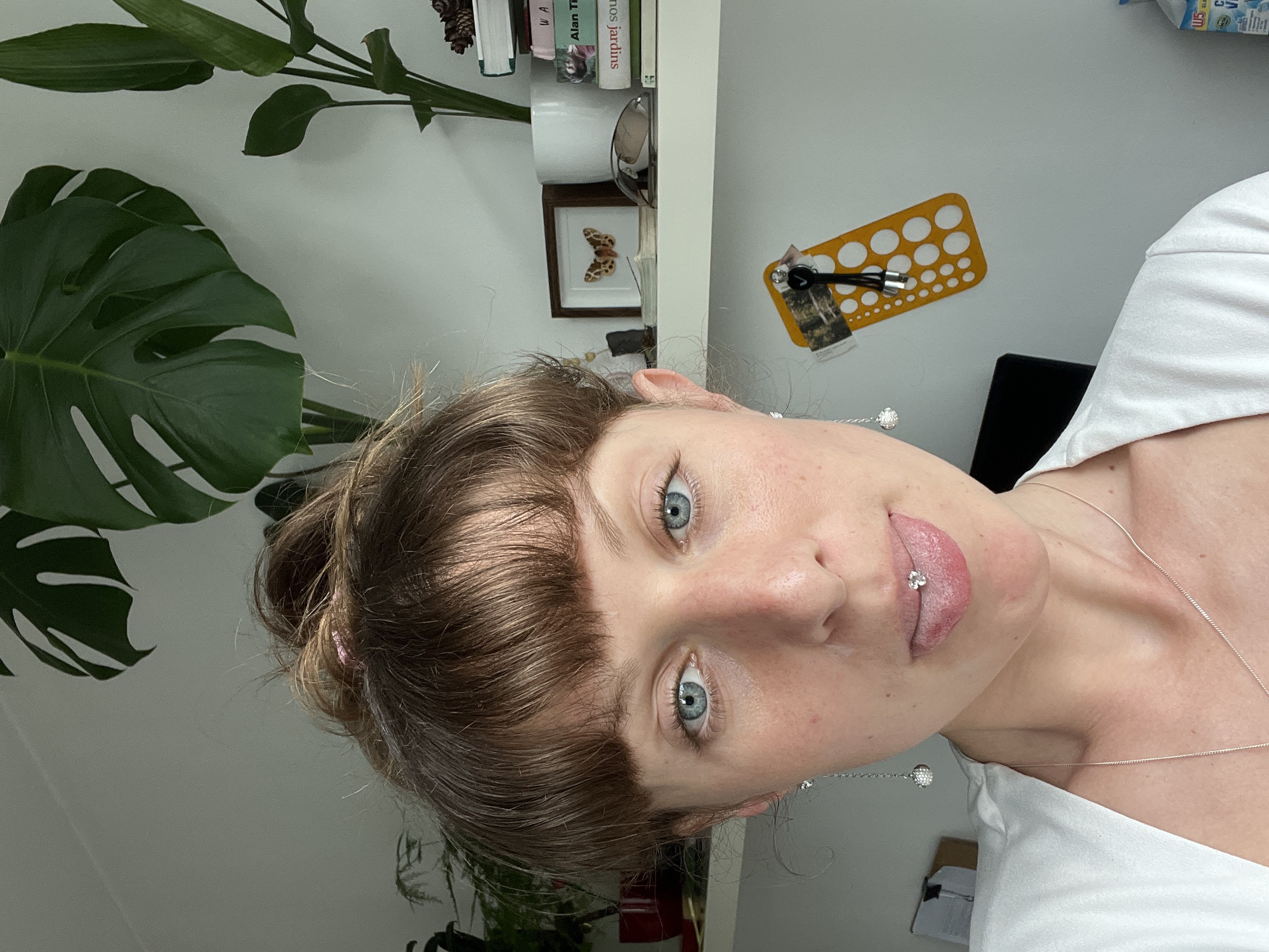 A close-up, high-angle shot features a woman with light skin, blue eyes, and light brown hair with bangs, a portion of her tongue visible with a piercing. She is wearing a white top and a silver necklace with a small spherical pendant. The background includes a white wall with a yellow stencil, a framed picture of a butterfly, books on a shelf, and green plants. The setting appears to be indoors.
