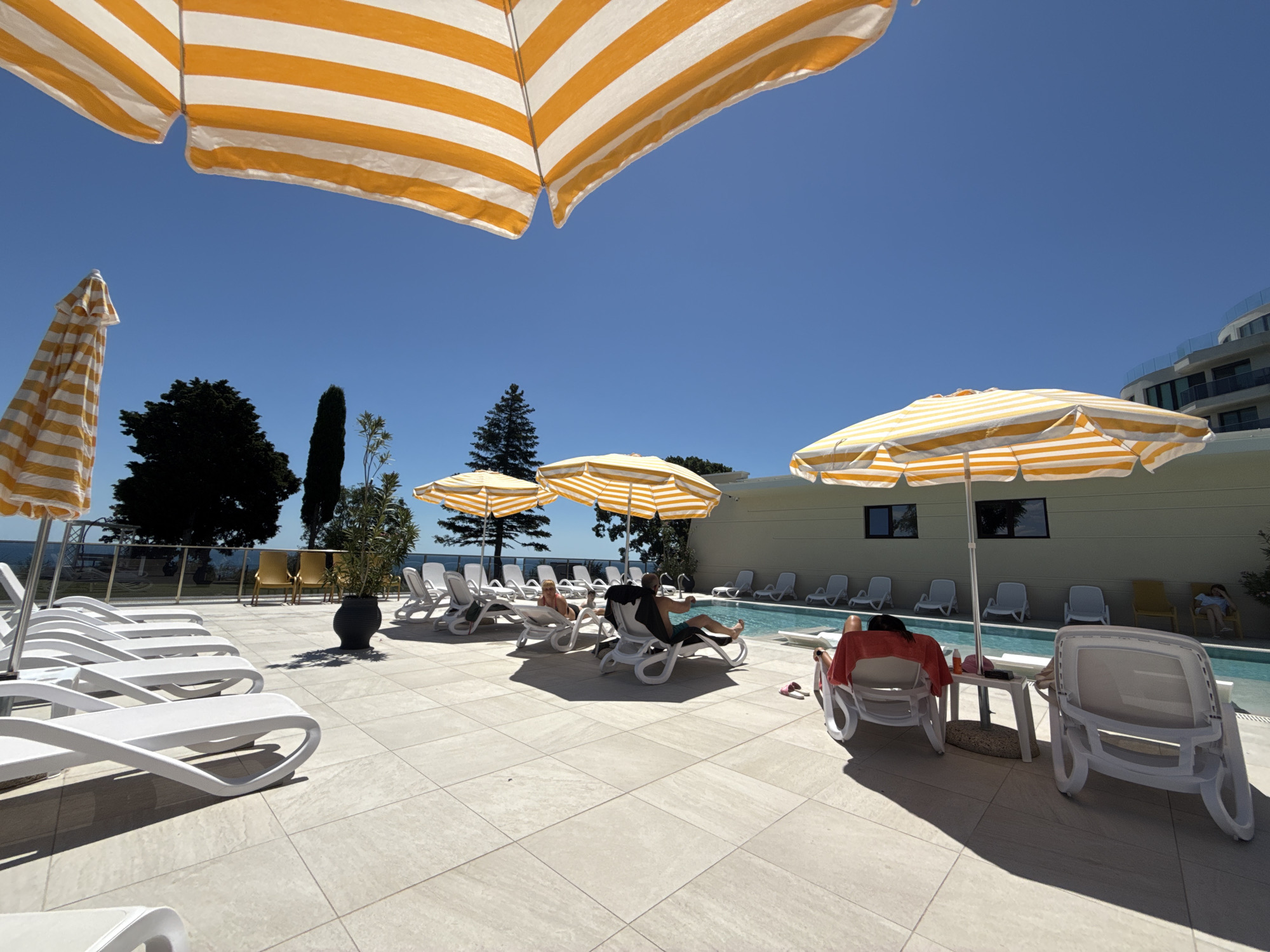 An outdoor swimming pool area is depicted under a clear blue sky. The ground is paved with light-colored square tiles. Several white lounge chairs are arranged on the patio, with some occupied by people. In the foreground, a row of empty white lounge chairs is visible on the left. Several orange and white striped umbrellas provide shade over some of the lounge chairs and a small table. In the middle ground, a few people are seen relaxing on lounge chairs. A swimming pool is situated behind some of the lounge chairs. In the background, a modern building with balconies and glass railings is visible, along with trees and a body of water that appears to be the sea. A black potted plant is placed on the patio.