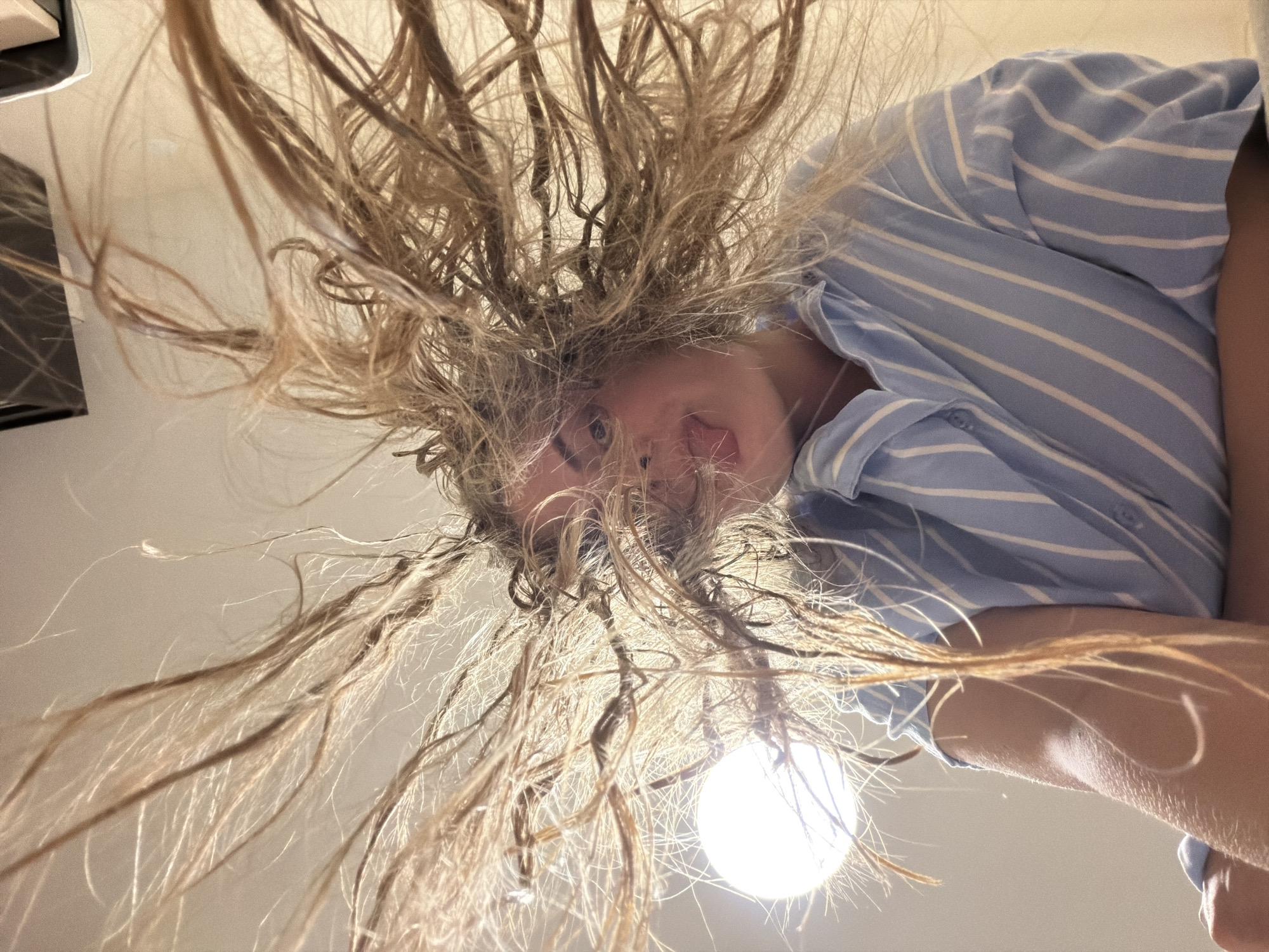 A person is positioned above the camera, facing downward. The person has blonde hair that is splayed outwards in a messy manner, partially obscuring their face. A light source, possibly a bulb, is visible below the person, creating an upward illumination. The person is wearing a blue and white striped shirt. The location cannot be determined.
