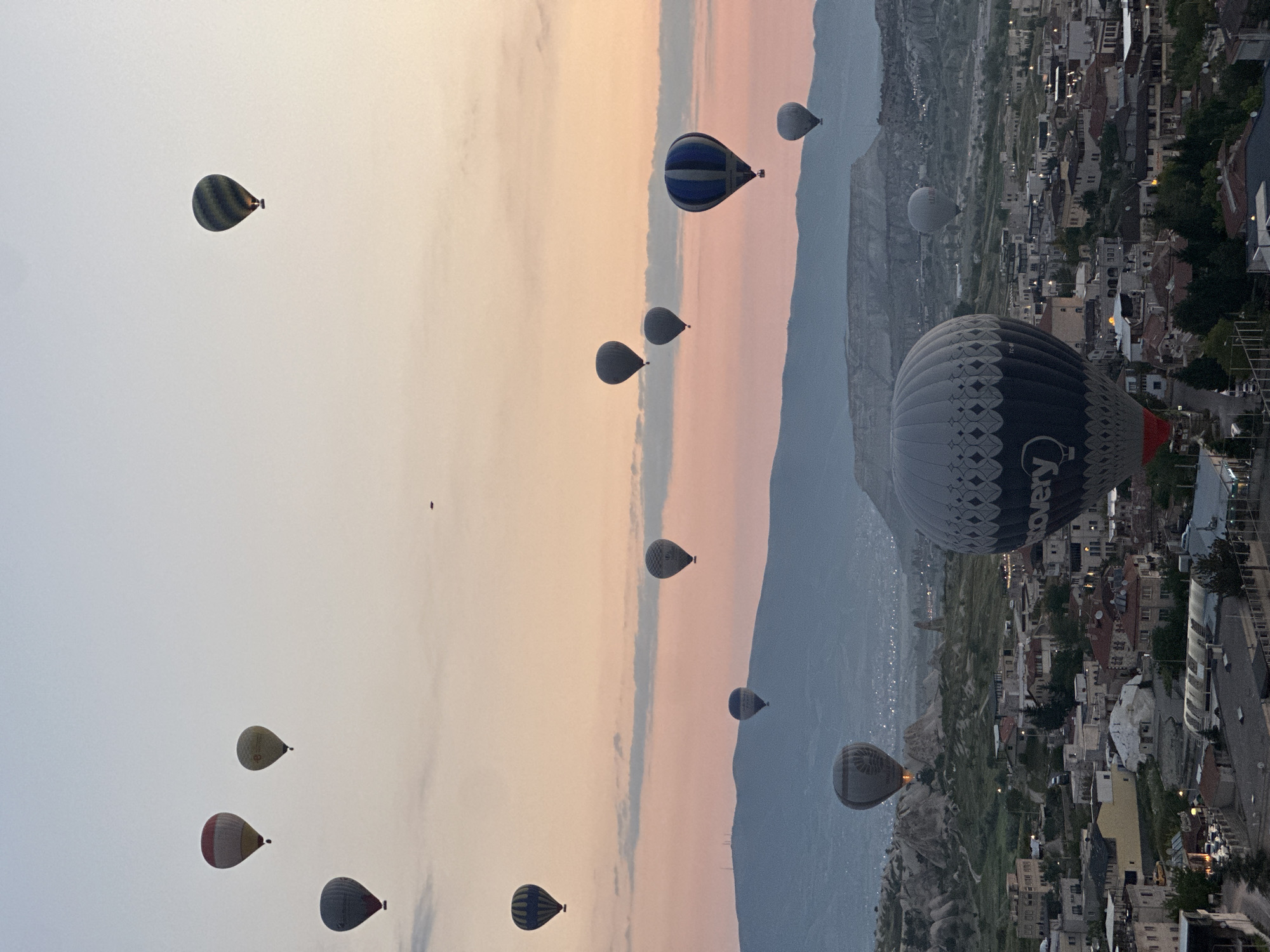 The image, when viewed in its correct landscape orientation, captures a serene and majestic scene of numerous hot air balloons in flight over Göreme, Turkey, during sunrise. The sky displays a beautiful gradient of colors, transitioning from a soft, light blue-grey at the top to warmer peach and pink hues near the horizon, typical of an early morning sky.
Across the vast expanse of the sky, approximately a dozen hot air balloons are visible, suspended at various altitudes. In the right foreground, a large, dark blue and grey balloon with an intricate patterned design is prominent. The text "Discovery" and a partial registration number "TC-..." are clearly visible on its envelope. Other balloons are scattered throughout the mid-ground and background, appearing as smaller, dark silhouettes or showing subtle patterns and colors like stripes or solid blue.
Below the balloons, a panoramic view of the unique Cappadocian landscape unfolds. Distinctive rock formations and valleys are visible, typical of the region. A town, likely Göreme, is nestled within this landscape, featuring numerous buildings and houses. Some artificial lights are still visible in the town, reinforcing the impression of dawn or pre-dawn activity. The overall scene evokes a sense of tranquility and awe, showcasing the iconic hot air balloon experience in Cappadocia.