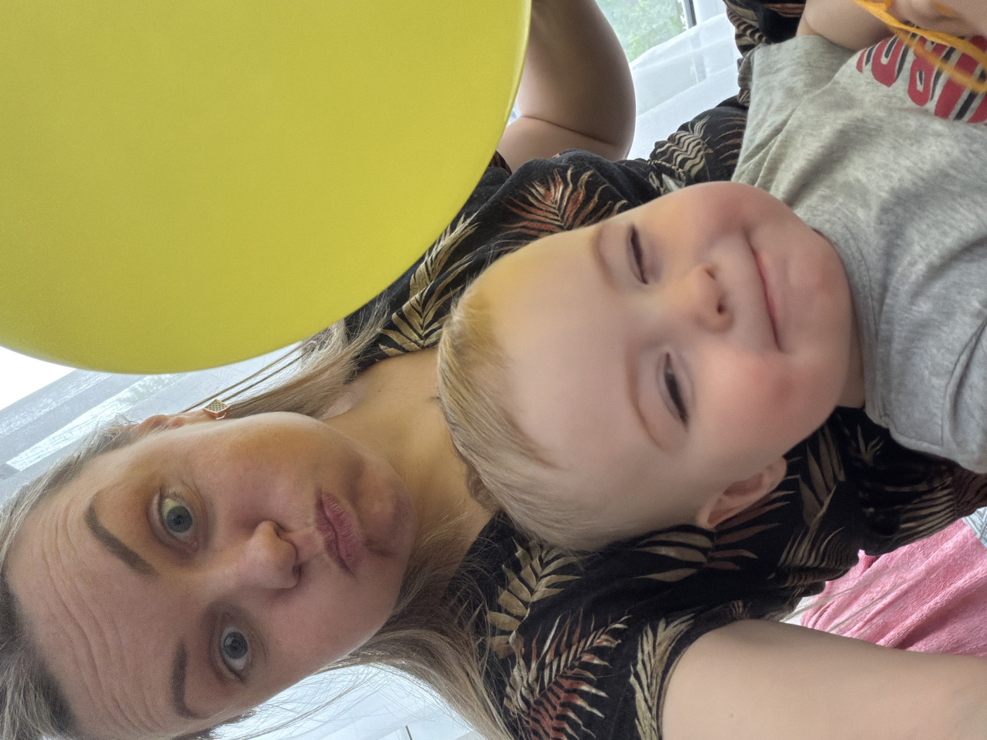 A selfie captures a woman and a child, likely taken indoors. The woman, fair-skinned with blonde hair, is making a puckered expression. The child, positioned beside her, is fair-skinned with blonde hair and a slight smile. A yellow balloon occupies the upper left of the frame. The woman wears a black floral-print top. The child is wearing a grey top with a red and white design. The background shows a blurred window. There is no information available that would establish this photo being taken in Votkinsk, Russia.