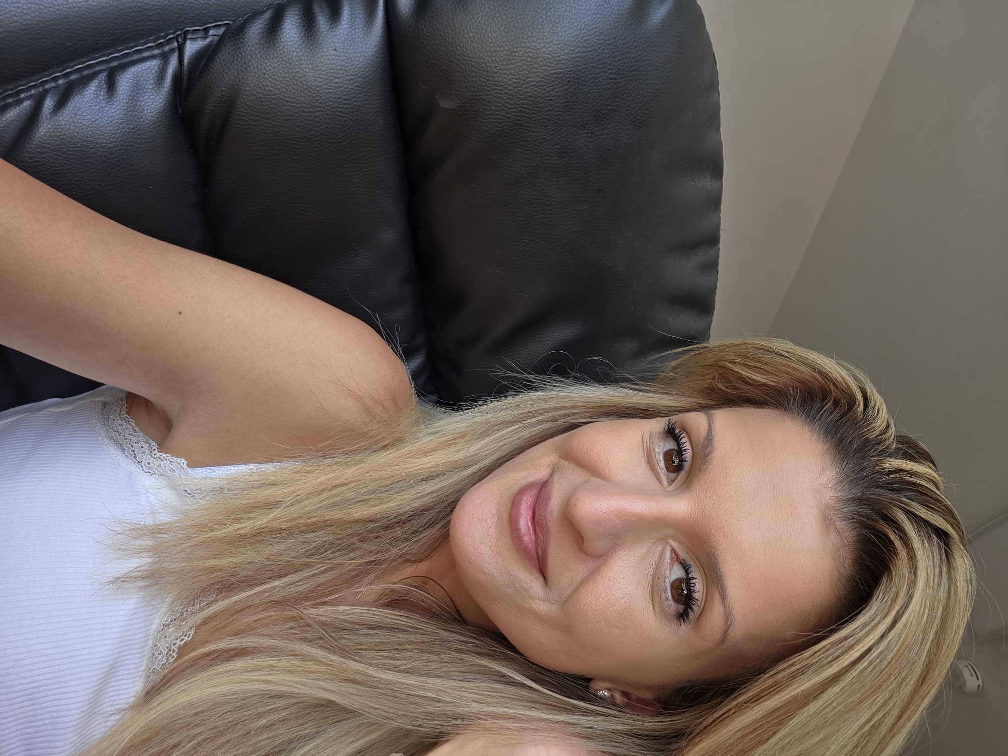 A medium shot captures a woman with blonde hair, seated in a black leather office chair. She is looking directly at the camera with a subtle smile. The background consists of a lightly colored wall, and part of a ceiling with a smoke detector is visible in the upper right corner. The woman is wearing a white tank top with lace trim. The scene appears to be indoors, likely an office or home environment. No specific location within Munich, Germany can be determined from the image.