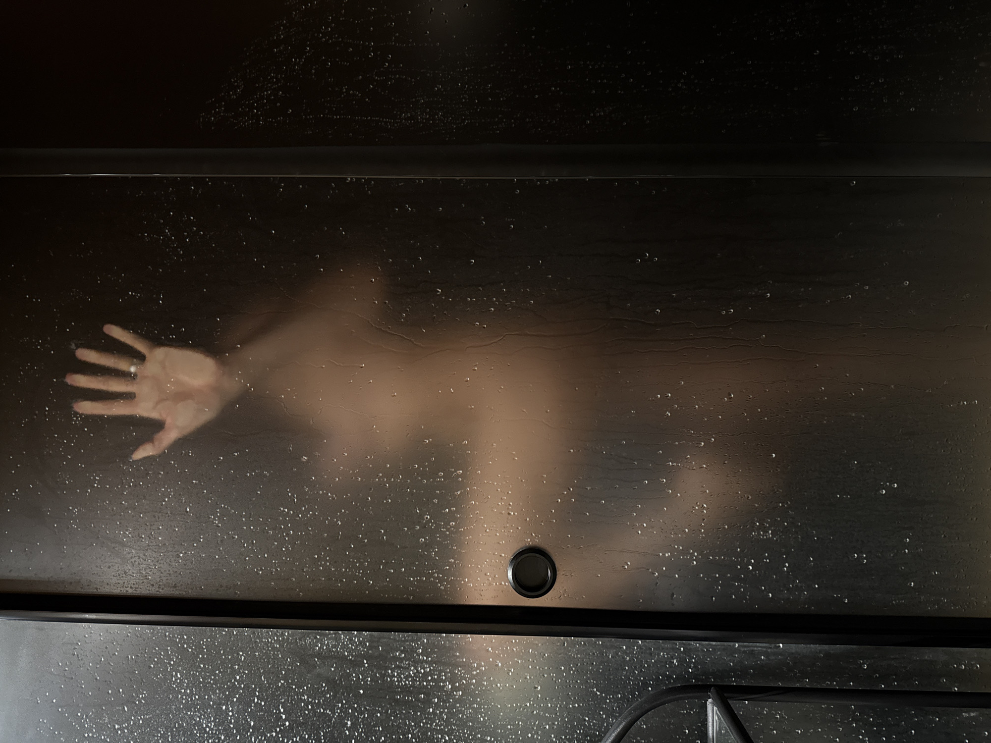 The image shows a blurred view of a person's upper body and hand pressed against a frosted glass shower door. Water droplets are visible on the glass. The person appears to be in a shower enclosure, with a dark background visible through the glass. A black handle is present on the door. The image was captured at an unknown location in the city of Chaikovsky, Russia.