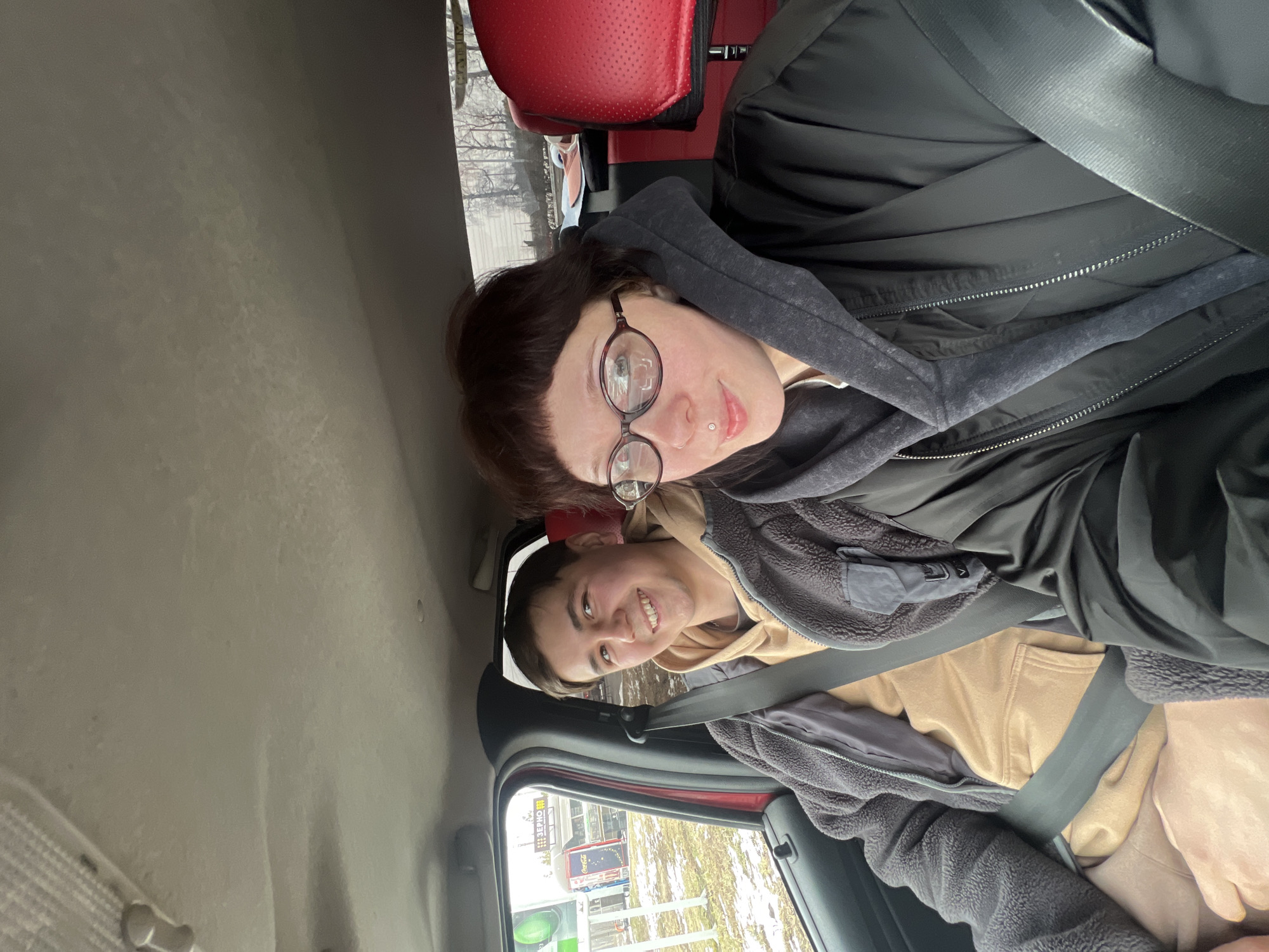 Two people are sitting in a car. One person is wearing glasses and a black jacket with a grey hoodie. The other person is wearing a tan hoodie with a grey jacket. Both are wearing seatbelts. Through the car window, there is a street scene in Kamennoye, Russia, with buildings and a Coca-Cola advertisement visible.