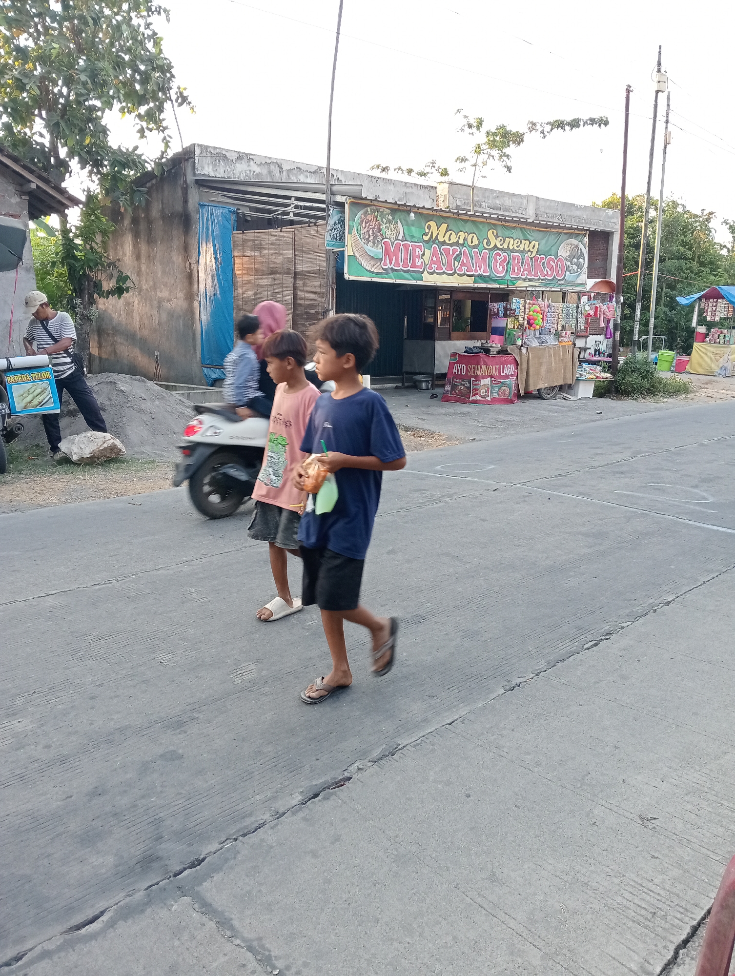 The image depicts a street scene in Tanjunganyar, Indonesia, characterized by a roadside establishment selling food, specifically "MIE AYAM & BAKSO." Two young boys are walking on the paved road. The boy on the left wears a pink t-shirt and dark shorts, while the boy on the right wears a blue t-shirt and black shorts. Both are barefoot or wearing flip-flops. In the background, a man in a striped shirt and cap is visible standing near a cart with a sign that reads "PAPEDA & IKOS." A motorcycle is parked nearby. The establishment features a prominent green banner with the text "Moro Seneng MIE AYAM & BAKSO" and displays various food items and promotional materials. The environment suggests a warm, possibly sunny day with trees and buildings lining the street. The road has clear white markings, and there are some cracks and debris on the surface.