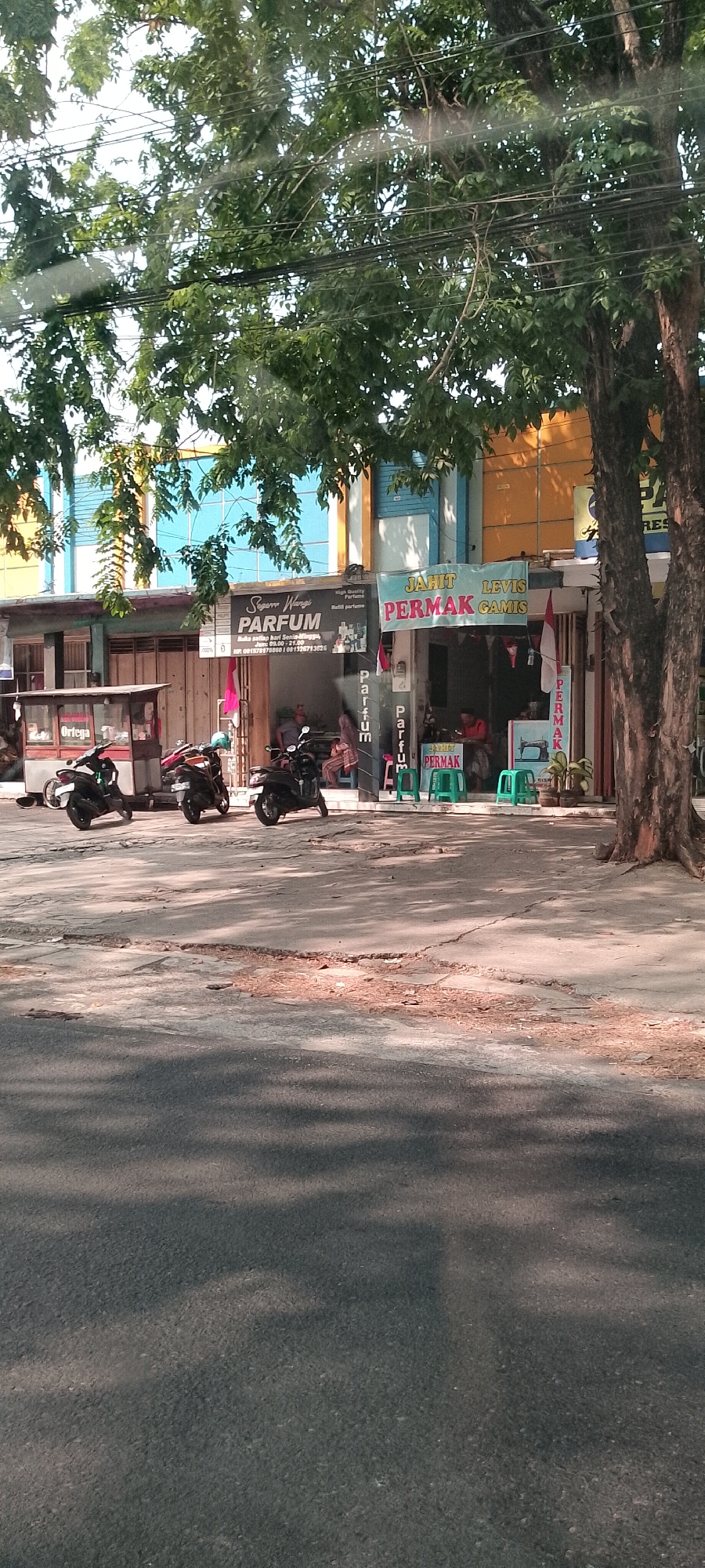 This image depicts a street scene in front of several shops in Panjang, Indonesia, under the shade of a large tree. The shops include a perfume shop named "Segarr Wangi PARFUM" which also lists its operating hours, and a tailoring/alteration shop named "JAHIT PERMAK GAMIS". Several motorbikes are parked in front of the shops. In the tailoring shop, there are people visible, one of whom appears to be working at a sewing machine, and green stools are placed outside. The street itself is paved with asphalt and shows some wear and tear. The overall impression is of a typical commercial area in Indonesia on a sunny day.