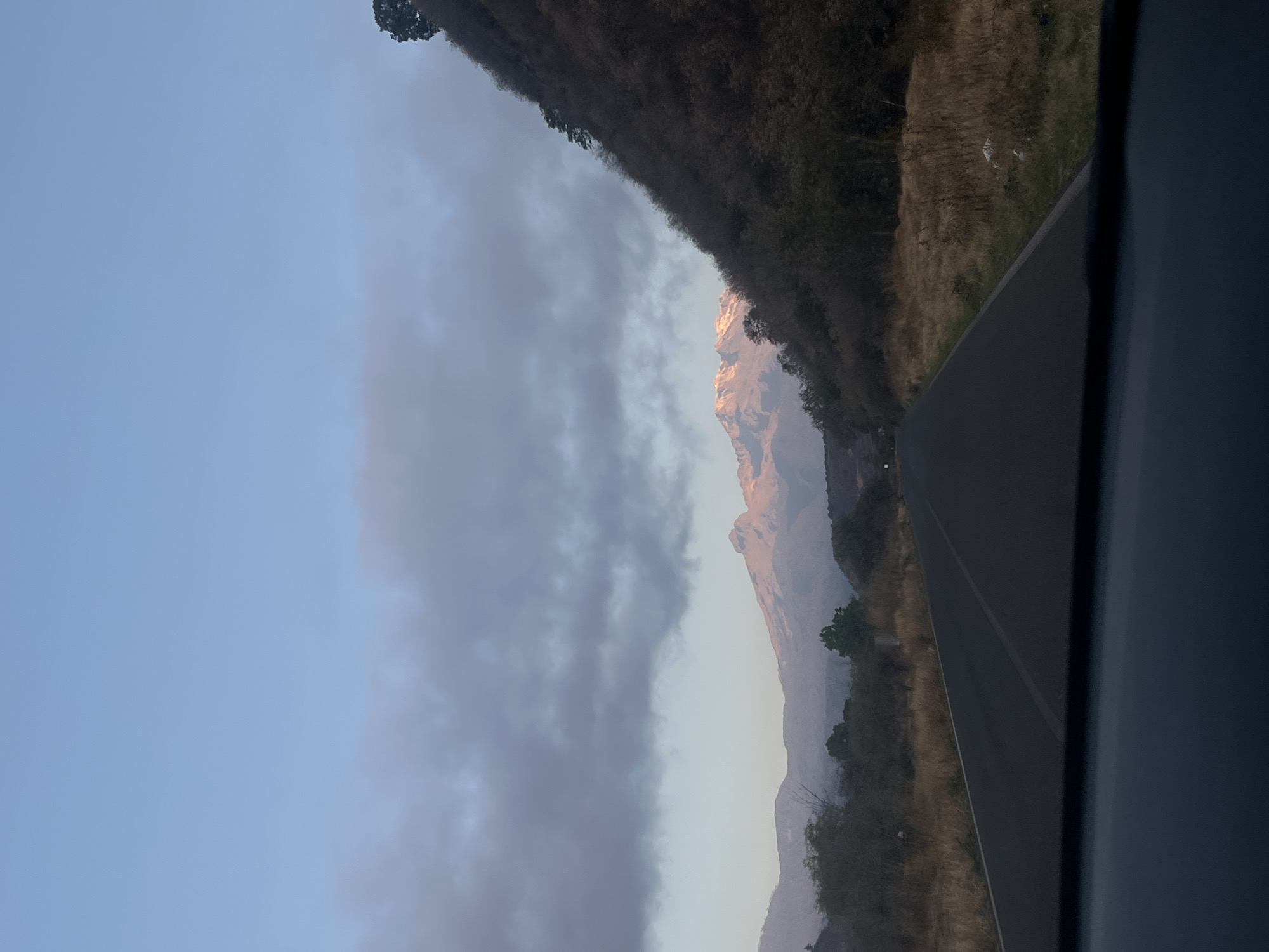 The image shows a mountain range, possibly near Santiago Xalitzintla, Mexico, partially obscured by clouds. The sky is light blue transitioning into the grey of the clouds. A road runs along the right side of the frame. Vegetation is visible along the roadside. The photo appears to have been taken from inside a vehicle, as a portion of the vehicle's interior is visible on the right. The mountain peak is highlighted by sunlight.