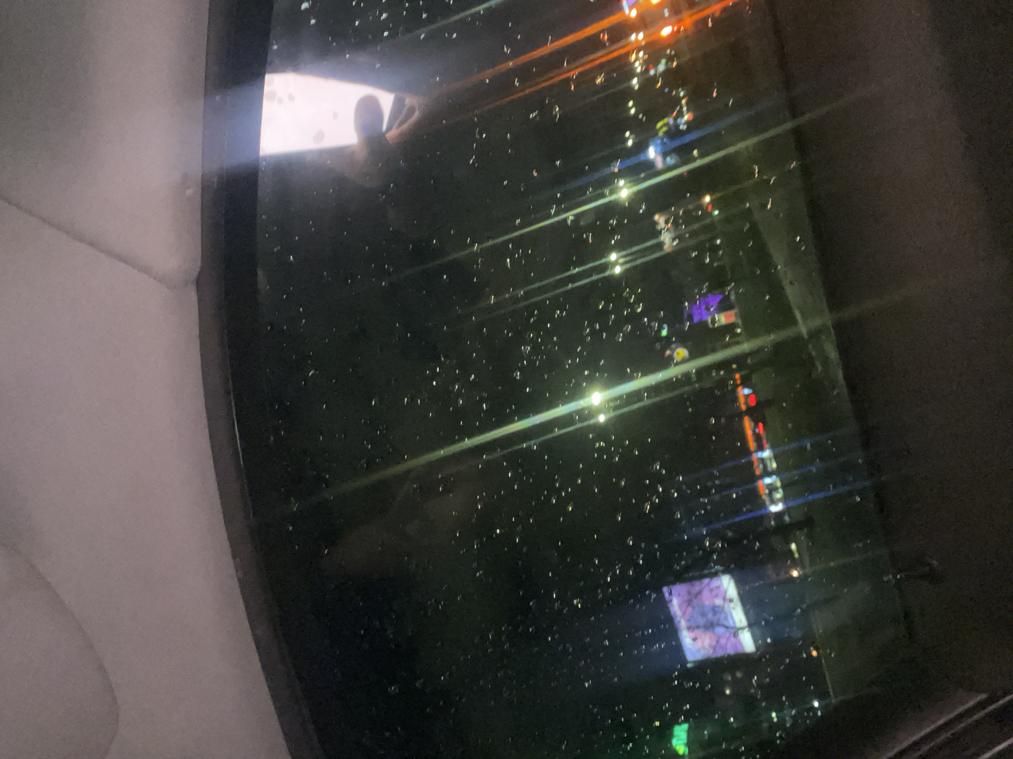 The image appears to be taken from inside a vehicle, possibly a car or bus, looking out the window. The window is covered in water droplets, suggesting it's raining or has recently rained. Through the window, blurred lights of the cityscape are visible, with streaks of orange, blue, and green indicating the presence of streetlights and illuminated signs. The reflection of a person is visible in the top left corner of the window, they seem to be seated. The general darkness suggests that the image was taken at night. I don't have information about the specific location within Sofia, Bulgaria.
