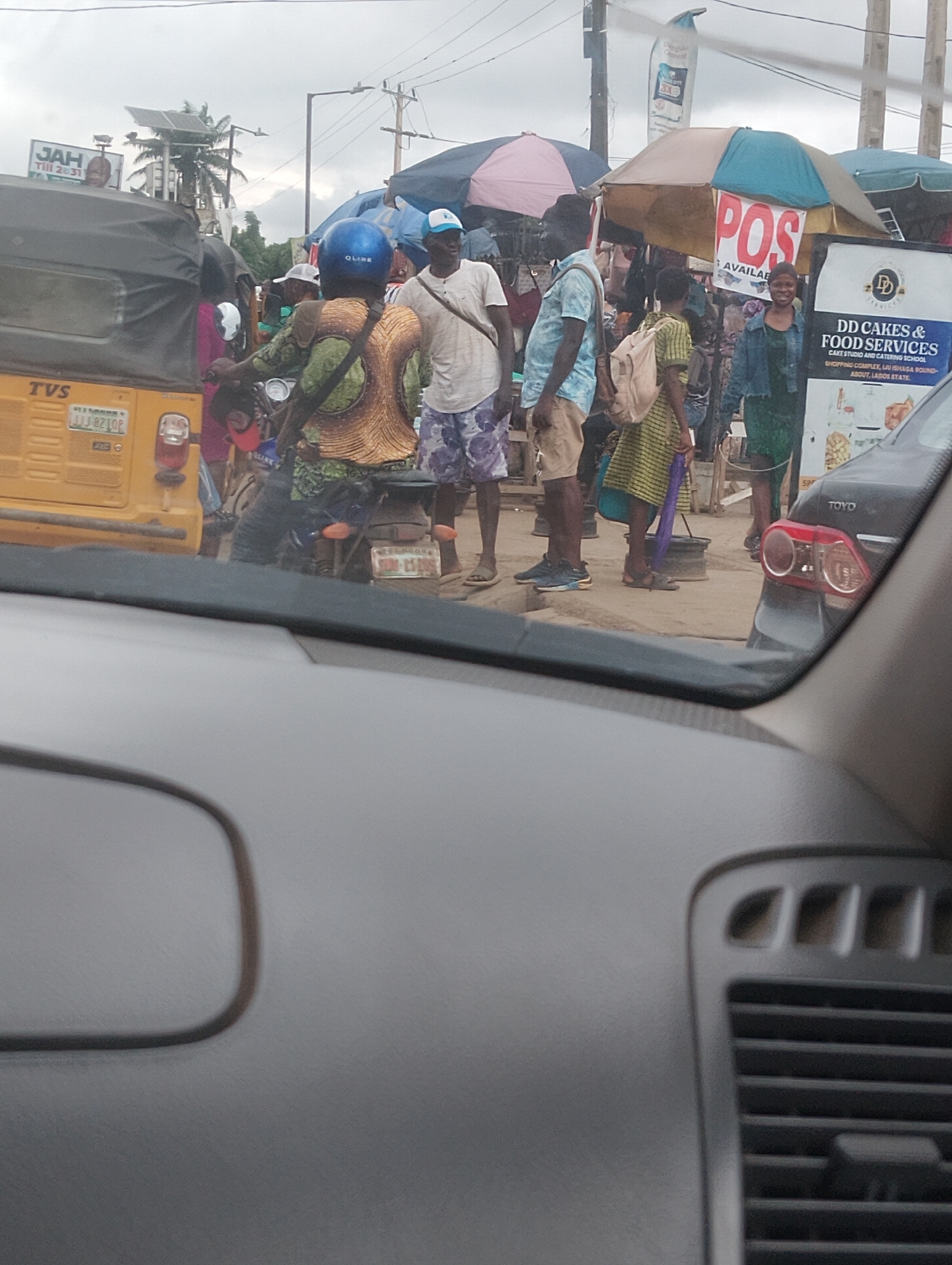 The image captures a street scene in Iju, Nigeria, on a cloudy day. In the foreground, the dashboard of a vehicle is visible. The middle ground features a bustling market with various vendors and shoppers. A yellow auto-rickshaw with "TVS" branding is parked on the left. Several people are interacting, some on motorcycles and others on foot. A woman in a green dress stands near a sign for "DD Cakes & Food Services". The background shows more market stalls with umbrellas and an overcast sky. The overall impression is one of everyday commerce and activity in a Nigerian urban setting.