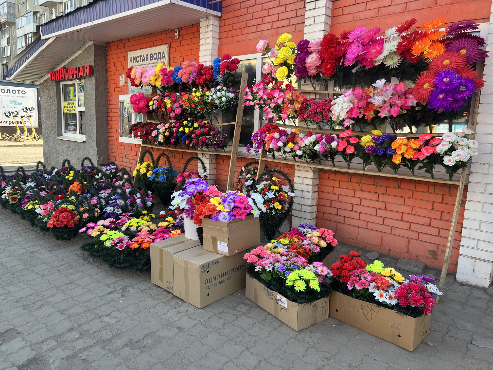 The scene is an outdoor flower shop located in Ufa, Russia, based on the text in the image. The shop sells artificial flowers and wreaths, displayed on shelves against a red brick building. The wreaths are arranged along the sidewalk. A sign that says "Чистая Вода" in Russian. Several cardboard boxes filled with artificial flowers. In the background, scooters are visible in the left of the frame.