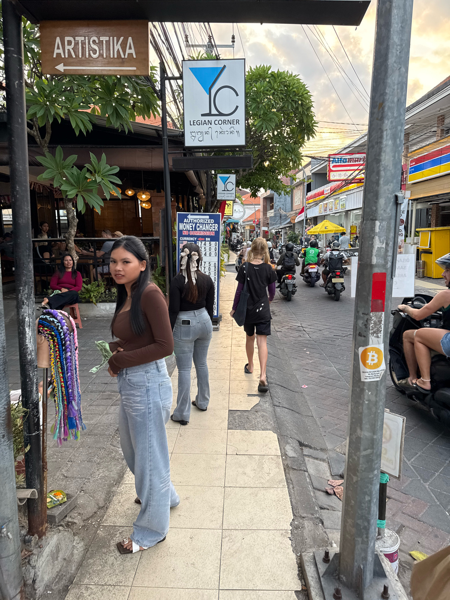 The image depicts a street scene in Kuta, Indonesia, likely during the late afternoon, with a warm, diffused light suggesting an approaching sunset. The foreground features a sidewalk lined with shops and a street on the right.
In the immediate foreground on the left, a young woman with long, dark hair is walking towards the camera. She is wearing a brown long-sleeved top and light blue wide-leg jeans. She holds some money in her right hand. To her left, a display of colorful braided hair extensions is visible, attached to a stand.
Further down the sidewalk on the left, other people are seated at tables outside a restaurant, partially obscured by foliage. On the right side of the street, a series of shops are visible, with awnings and signs, including an "Alfamart" convenience store. Several motorbikes are on the street, some with riders wearing helmets. One motorbike in the mid-ground has a rider in a green jacket.
A tall, grey metal pole stands prominently on the right side of the image, displaying stickers, one of which is a Bitcoin logo. The street is paved with cobblestones. Overhead, a tangle of electrical wires is visible against a sky with scattered clouds.
A signpost is present on the left, with a wooden sign labeled "ARTISTIKA" and an arrow pointing left. Another signpost further back displays "LEGION CORNER" with some script below it, and a smaller sign below that indicates "AUTHORIZED MONEY CHANGER". The overall impression is of a busy commercial street in a tropical urban environment.