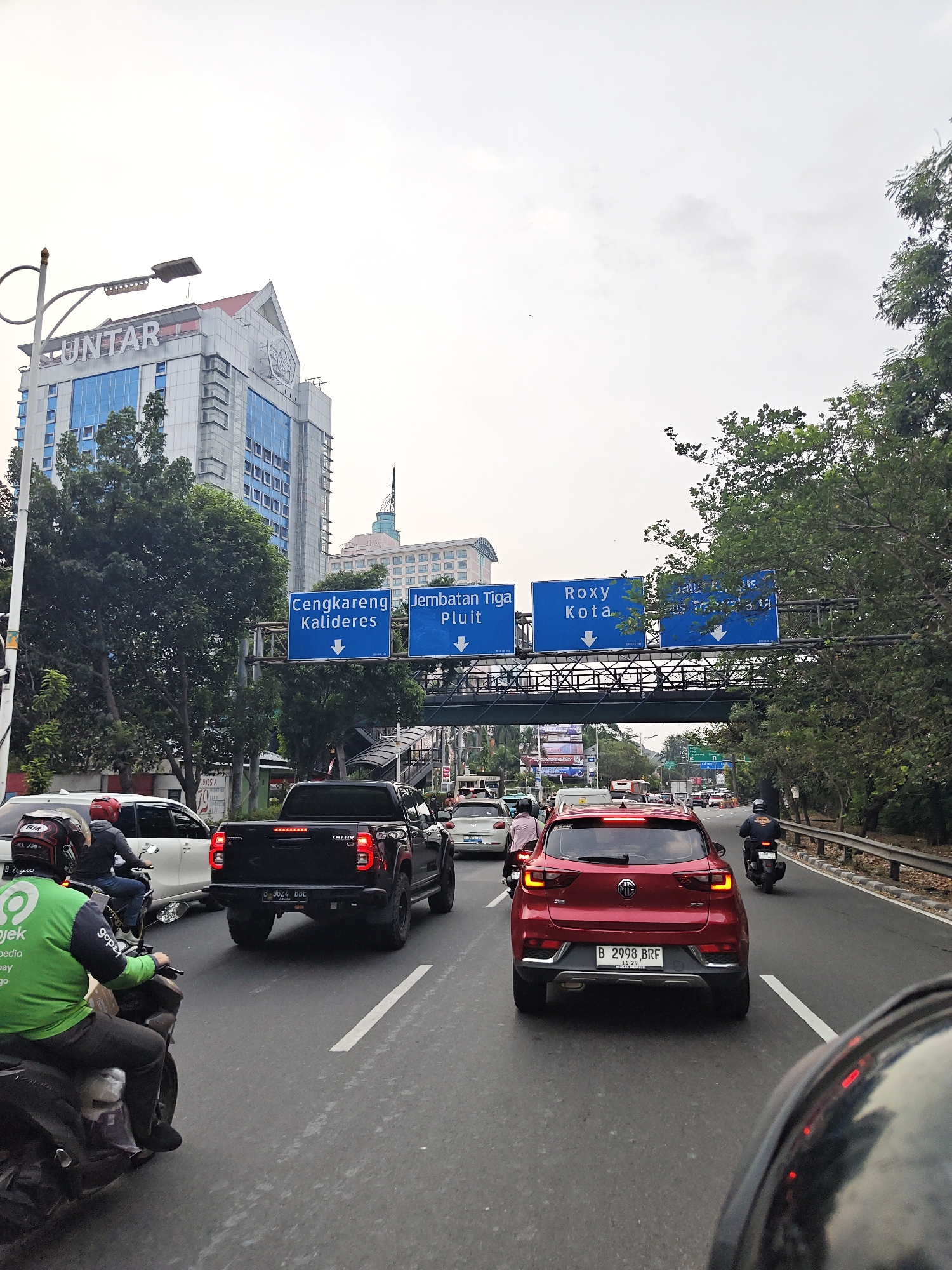 The image shows a traffic scene on a road in West Jakarta, Indonesia. Multiple vehicles are present, including cars, a pickup truck, and motorcycles. A rider in a green Gojek uniform is visible on the left side of the image, on a motorcycle. Road signs indicate directions to "Cengkareng Kalideres," "Jembatan Tiga Pluit," and "Roxy Kota." In the background, there are several buildings, including a tall, modern-looking building with the word "UNTAR" on it, and other office buildings. Trees line the right side of the road. The sky is overcast. The perspective suggests the image was taken from a vehicle in traffic.