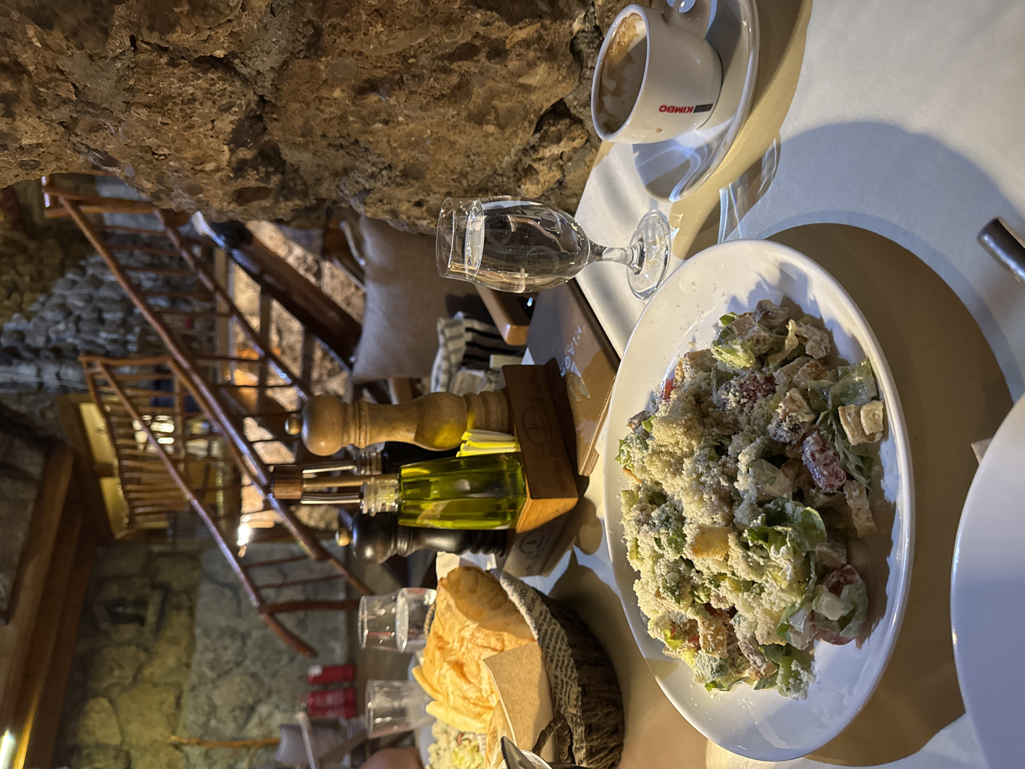 The image depicts a table setting in a restaurant, likely located within a cave or stone structure, given the visible rough stone walls and ceiling. Wooden stairs can be seen in the background. On the table, there is a white plate with a salad topped with grated cheese, a glass of water, a cup of coffee with "Kimbo" branding, a wooden stand holding a pepper grinder and a bottle of olive oil, and a basket containing bread. The setting suggests a restaurant in Podgorica, Montenegro, potentially within a unique, natural environment, but no people are clearly visible.