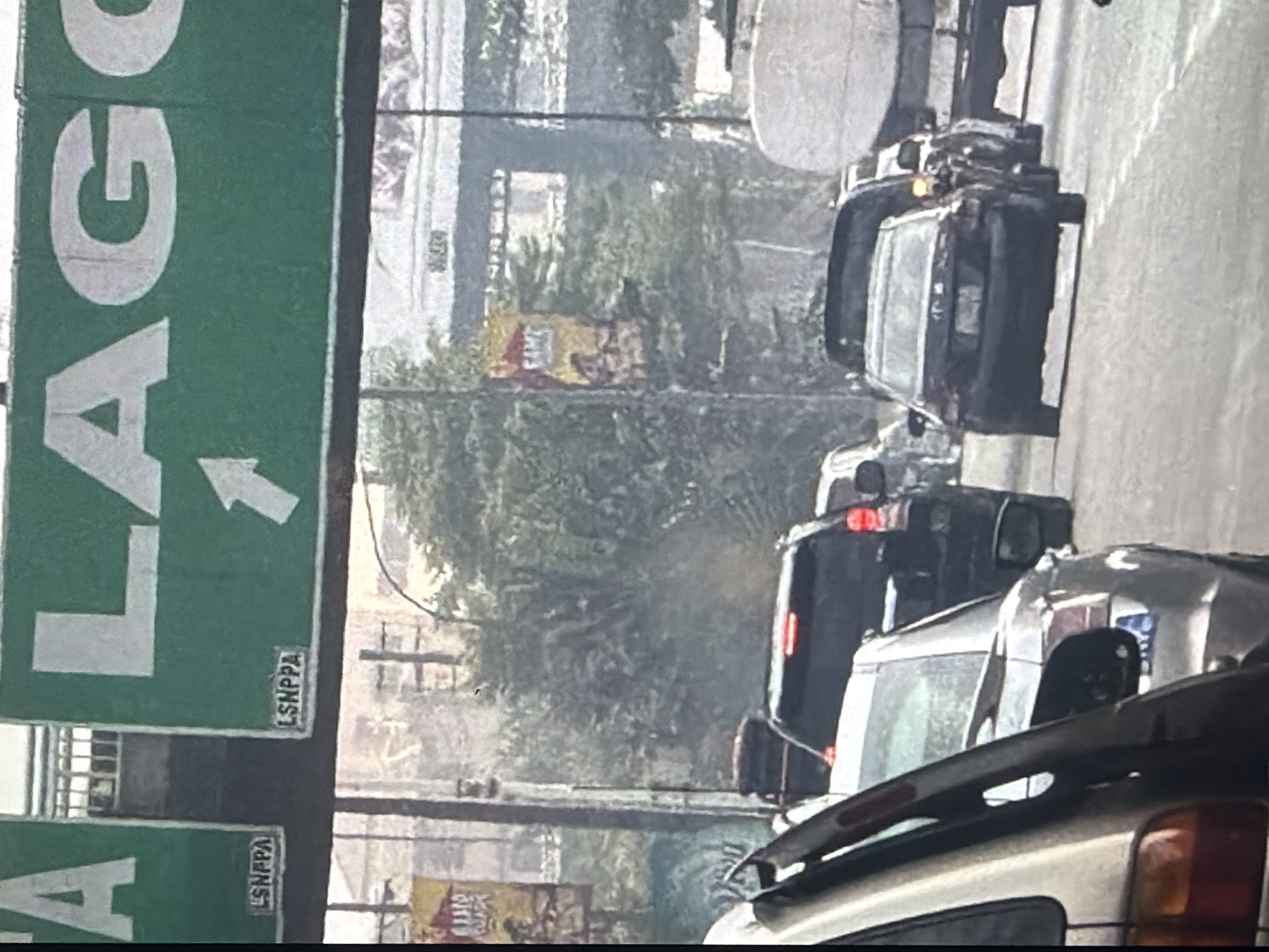 The image shows a traffic jam on a highway. A green sign with white lettering is visible at the top, with the word "LAGOS" and an arrow pointing right. Below "LAGOS" are two smaller signs that read "LSNPPA". There are several cars on the road, including a black SUV, a silver sedan, and a black sedan. A large truck is also visible in the background. Trees and buildings can be seen in the distance. The image appears to be taken from a vehicle in traffic. The traffic jam is likely on the highway leading to Lagos, Nigeria, possibly in the city of Bukuru.