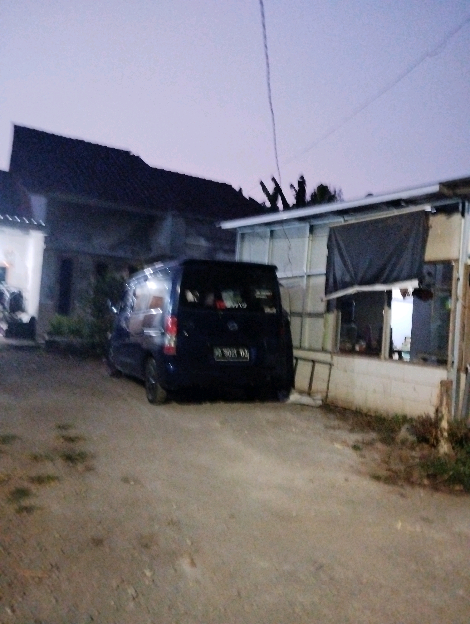 In Salatiga, Indonesia, an outdoor scene unfolds at dusk. The focal point is a dark blue van parked on a dirt and gravel surface. The van's license plate is partially visible, reading "AD 3021 BJ". Adjacent to the van is a structure with a corrugated metal roof and walls, partially covered by a dark tarp. A residential building with a dark tiled roof is visible to the left of the van. The sky is a pale, dusky purple, with faint outlines of power lines. The overall impression is a quiet, residential street scene at the end of the day.