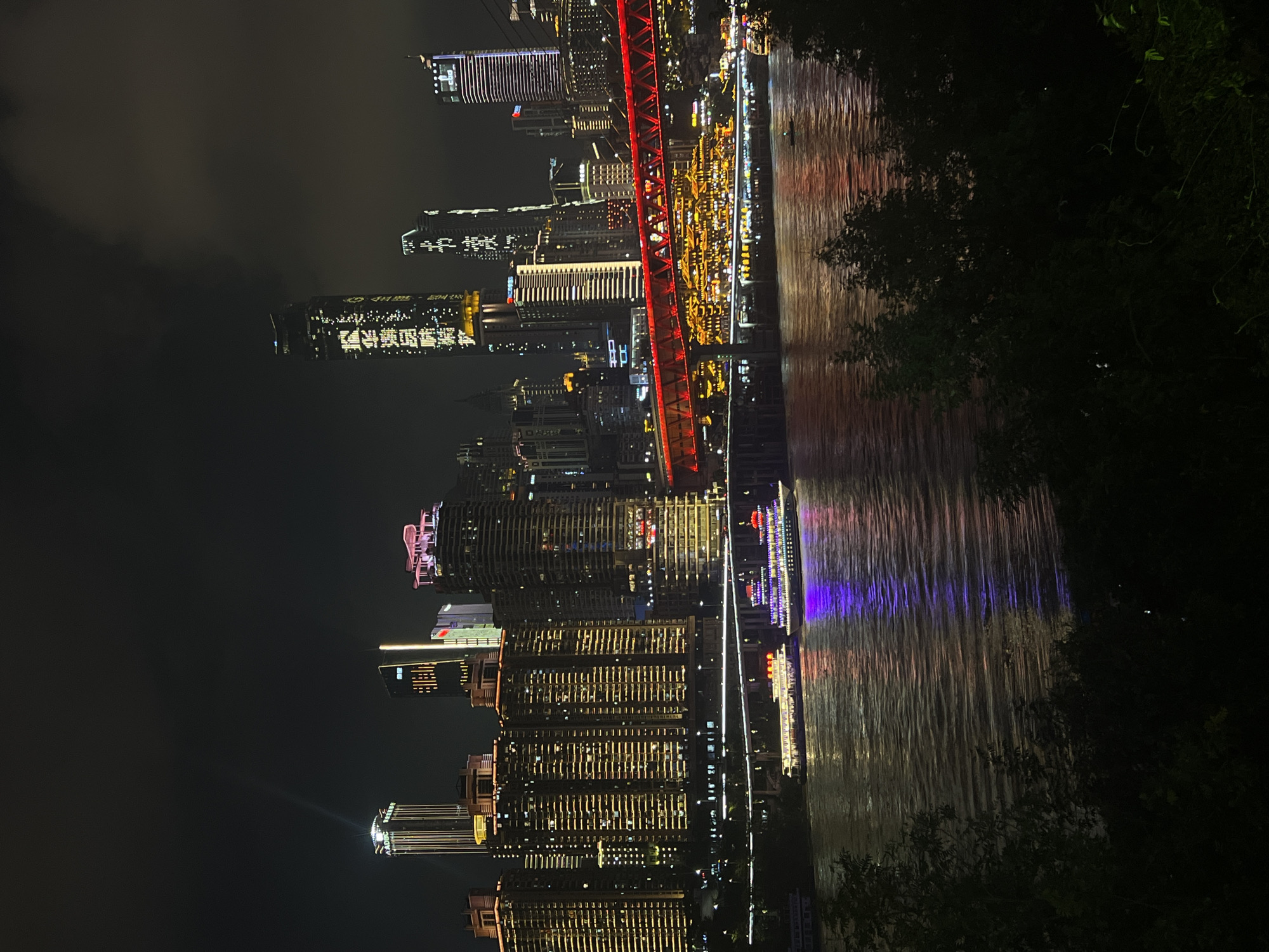 The image shows a nighttime view of the cityscape and a river in Jiangbeicheng Street, China. Numerous tall, illuminated buildings form a dense urban skyline across the river. A bright red illuminated bridge spans the river, with traffic visible on it. Below the bridge, two large passenger boats are docked or moving on the river, their lights reflecting on the water. The river itself reflects the colorful lights of the city and the boats. In the foreground, dark silhouettes of trees and foliage obscure the bottom portion of the image. The sky above is dark with some cloud cover.