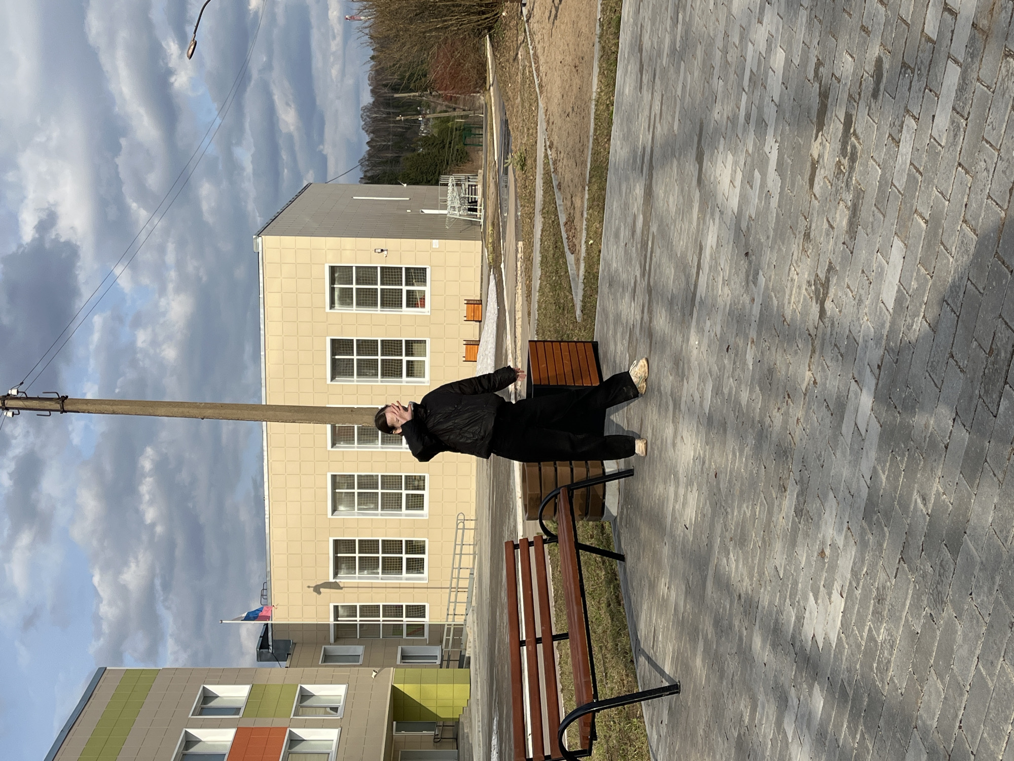 The image captures an outdoor scene in Сосново, Russia, featuring a person standing on a paved area amidst buildings under a cloudy sky.
In the midground, a person, likely an adult, is dressed in a dark (black) jacket, black pants, and light-colored sneakers. They are standing near a wooden bench with black metal supports, with their left hand raised towards their head or face, possibly shielding their eyes or adjusting something. Their back is slightly turned towards the camera.
The ground in the foreground and around the person is paved with grey rectangular bricks. Beyond this, a dirt or gravel path with sparse, dry grass patches is visible, leading towards a wooded area in the background with bare trees, suggesting a cooler season.
Several buildings populate the scene. A prominent, multi-story building with a light-beige or yellowish facade and numerous white-framed windows is situated in the mid-left. Below it, a section of another modern building with a grey roof and colored panels (green and orange) is partially visible. A small flag, displaying horizontal stripes of blue, pink, and white, is seen flying from a pole attached to the beige building.
A tall utility pole with overhead wires stands on the left side of the frame, and a street light fixture is visible near the top edge. The sky is overcast with broken clouds, but there's enough sunlight to cast distinct shadows from the benches and buildings onto the paved ground. No discernible text is visible in the image.