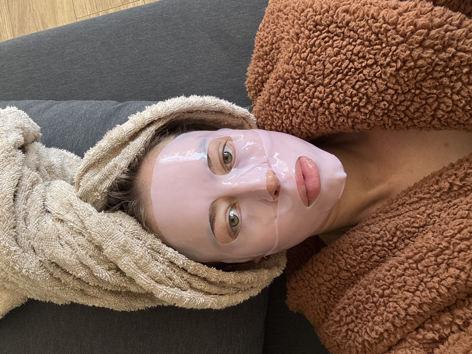 An individual is depicted in an indoor setting, likely a private residence, in the city of Unknown, Unknown. The subject is a female, seen from an overhead perspective, reclining. Her face is covered by a translucent, light pink sheet face mask with cutouts for the eyes, nostrils, and mouth. Her eyes, open and appearing green or hazel, look directly into the camera. A light brown terry cloth towel is wrapped around her head, covering her hair. She is wearing a textured, brown, fleece-like bathrobe or garment, visible over her upper torso. The immediate background consists of a dark grey upholstered surface, likely a sofa or bed. A light-colored wooden floor is visible in the upper right portion of the frame. No discernible interactions are present.