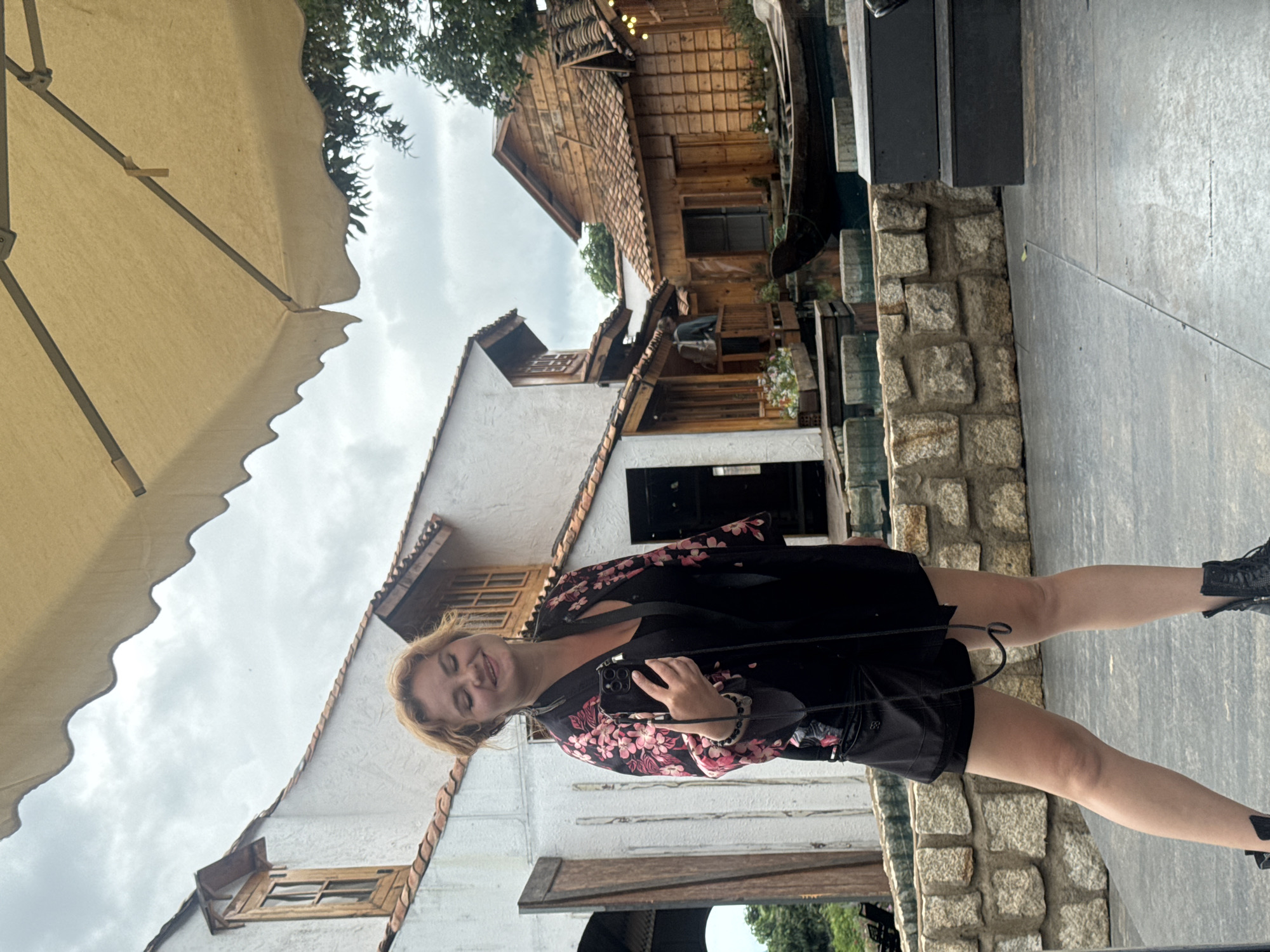 An overhead view shows a young Caucasian woman with blonde hair, wearing a black dress and a floral kimono, posing for a selfie with her phone. She is standing on a paved surface with a stone wall behind her. In the background, there are several traditional Vietnamese-style wooden houses and a small boat on a canal. The location is Thôn 3, Vietnam. The sky is overcast.