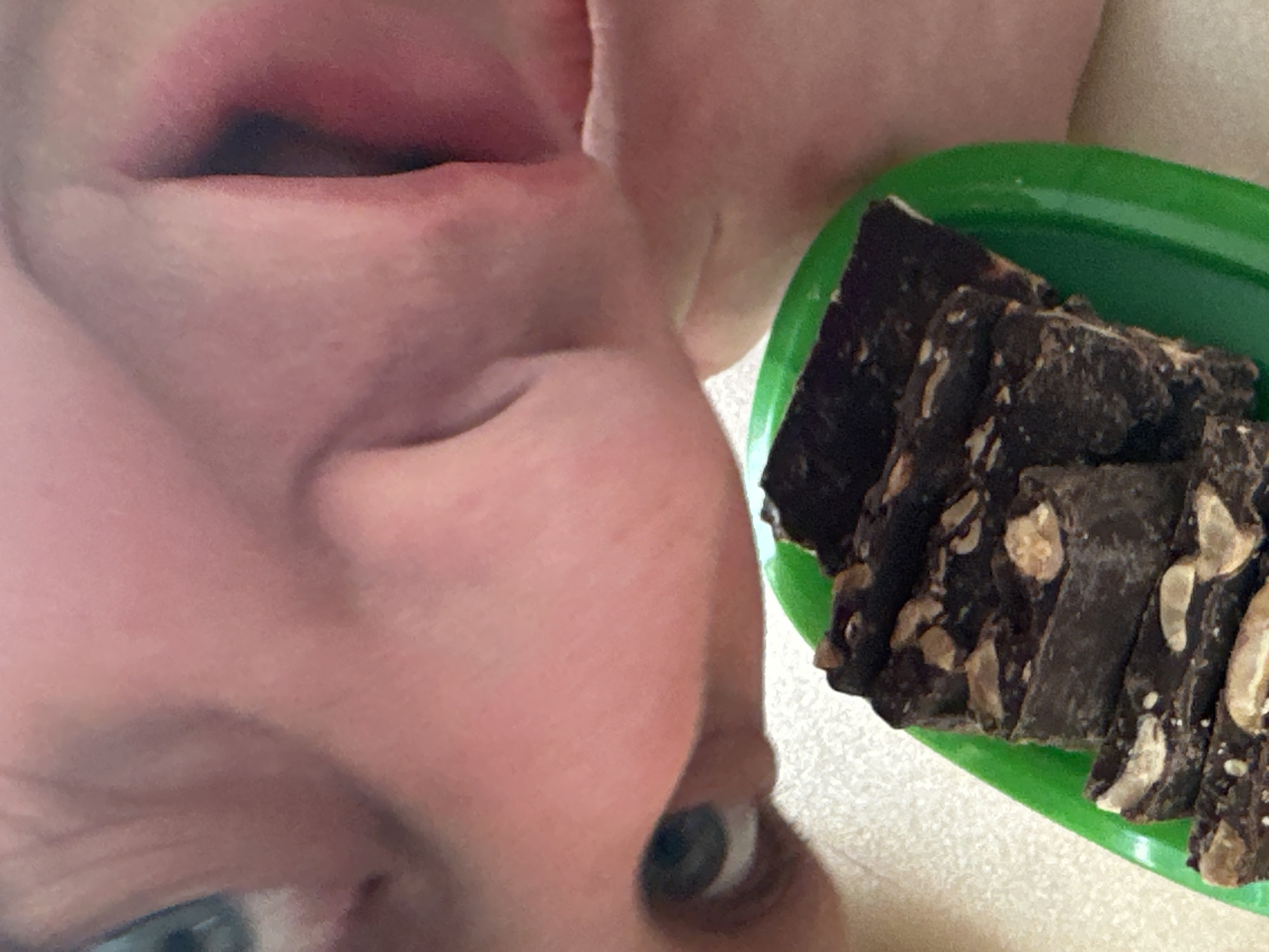 A close-up, slightly overhead photograph captures the lower portion of a human face and a food item. The upper half of the frame displays the left eye, nose, and an open mouth with a visible tongue belonging to an individual with fair skin and a blue iris. The gaze is directed downwards and to the right. Below the face, a bright green, oval-shaped plate or container holds a stack of approximately eight dark brown, rectangular food items, consistent with chocolate fudge or bark, interspersed with lighter-colored particulate matter, likely nuts. The food items are positioned on a light-colored, subtly textured surface, possibly a countertop or table, which forms the background. The individual's open mouth and downward focus indicate an imminent or ongoing interaction with the food. The image provides no discernible features to identify a specific location within the city of Chaikovsky, Russia.