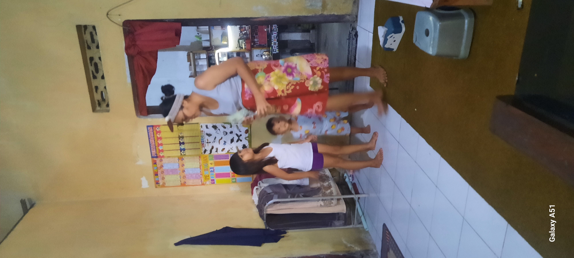 In an indoor residential setting in Ploso, Indonesia, a woman wearing a grey tank top and a floral skirt stands facing a young girl. The woman has dark hair pulled into a bun and wears glasses and a headband. The young girl, wearing a white tank top and purple shorts, stands to her left. To the right of the girl, a young child in a light blue outfit with a print of airplanes is looking towards the woman. The background features a yellow wall with educational posters, including one with insect illustrations and another titled "PENAMBAHAN" (addition) and "PEMBAGIAN" (division). A clothes rack with various garments is visible to the left of the girl. The floor is tiled in white. In the foreground, a brown rug is present with a tissue box and a small grey stool. A doorway with a red curtain is visible to the right of the woman. The overall impression is a casual indoor scene, possibly a domestic living area or classroom.