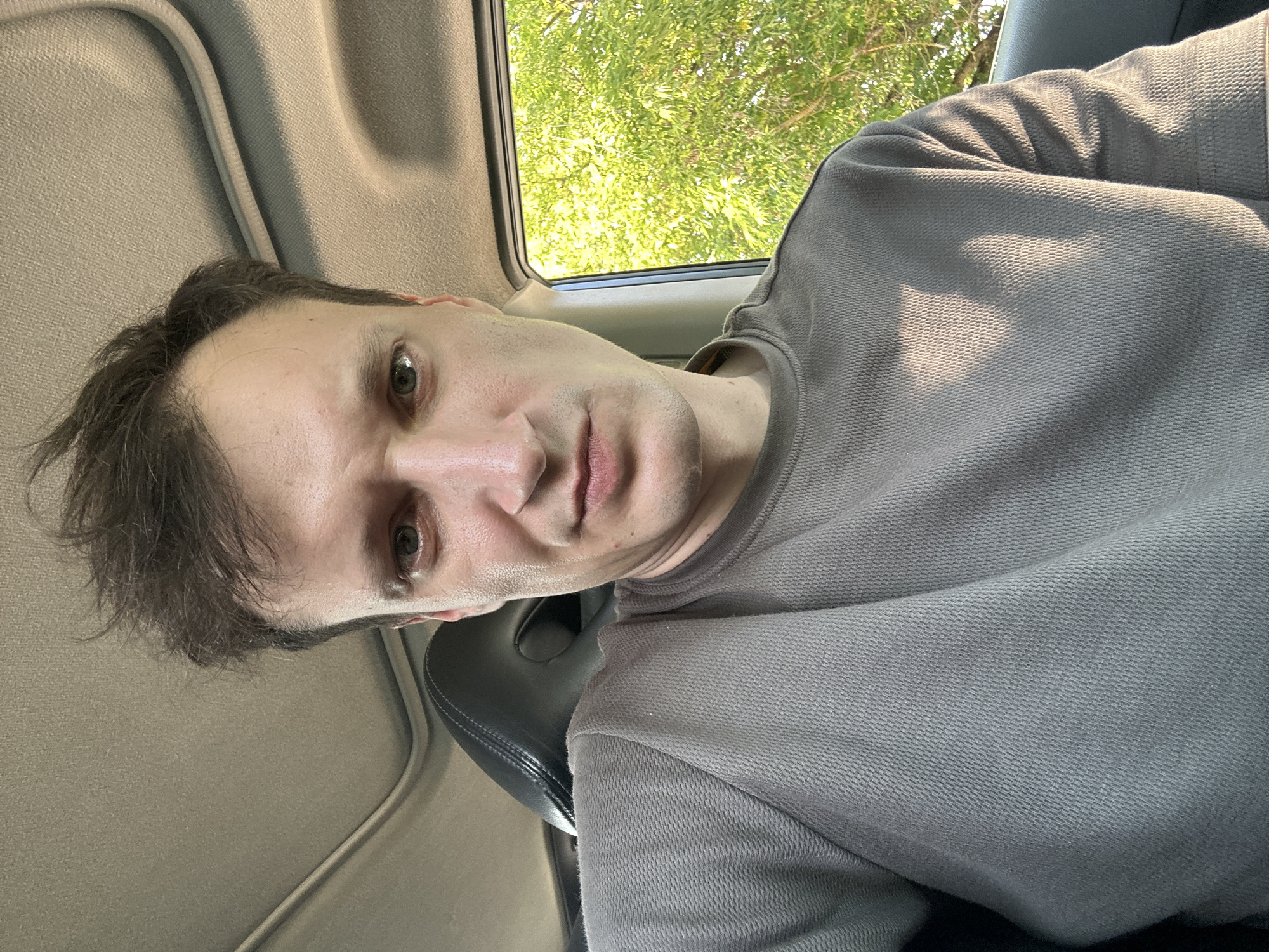 A close-up selfie taken from inside a car shows a Caucasian man with short, messy brown hair. He is wearing a dark gray long-sleeved shirt. The man is looking directly at the camera with a neutral expression. His right eye is visible, and his left eye is partially obscured. Sunlight is filtering through the trees outside the car window, casting dappled shadows on his face and shirt. The interior of the car, including the headrest and sunroof, is visible in the background. The scene appears to be in a natural setting, possibly a park or a wooded area, given the prominent greenery. There is no indication of the city of Makiv, Ukraine, in the image.