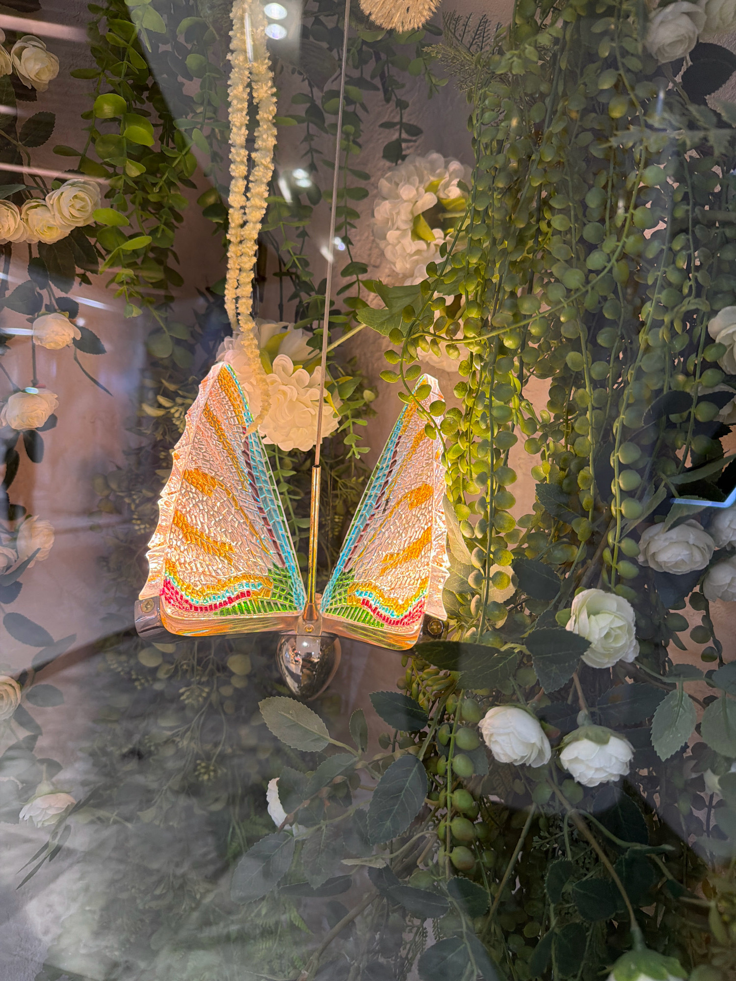 A decorative butterfly-shaped lamp is suspended amidst artificial greenery and flowers, likely displayed in a shop window. The lamp's wings are translucent with colored patterns resembling stained glass. The surrounding artificial foliage includes hanging vines with small green beads and clusters of white roses. The scene is viewed through a reflective surface, possibly glass, which casts distortions and reflections onto the display. The location is identified as Pavlodar, Kazakhstan, but no specific details about the city or its establishments are visible in the image.